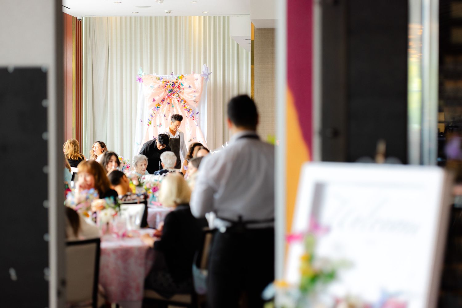 Elegant wedding reception with guests seated at decorated tables while a waiter serves in a softly lit venue.