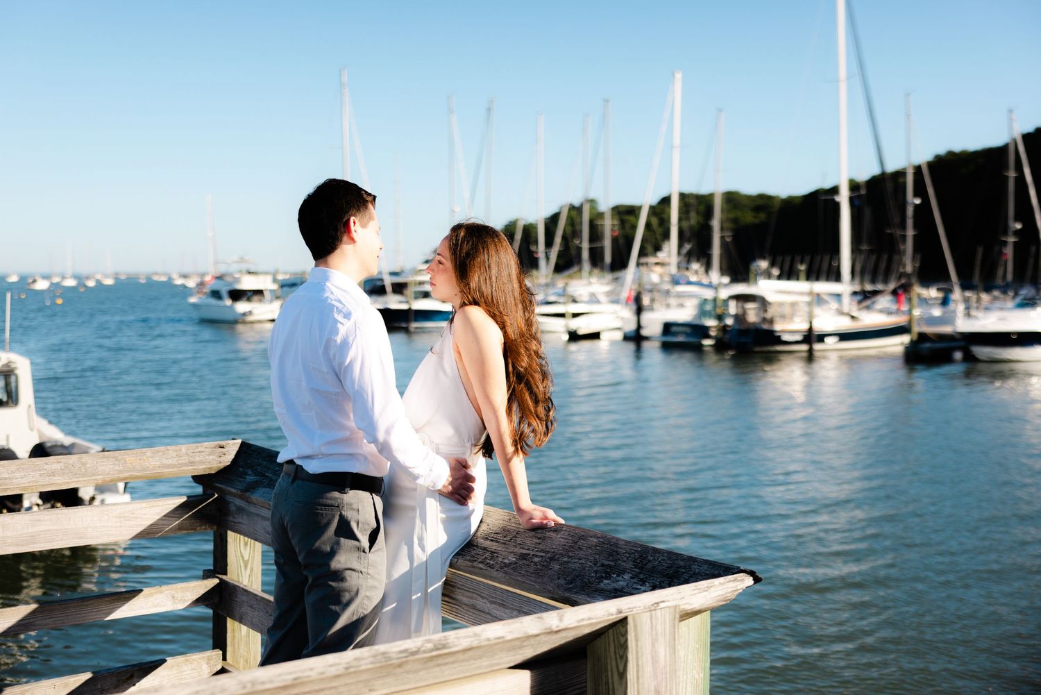 Couple in wedding attire standing on a wooden dock overlooking a marina with sailboats on a sunny day.