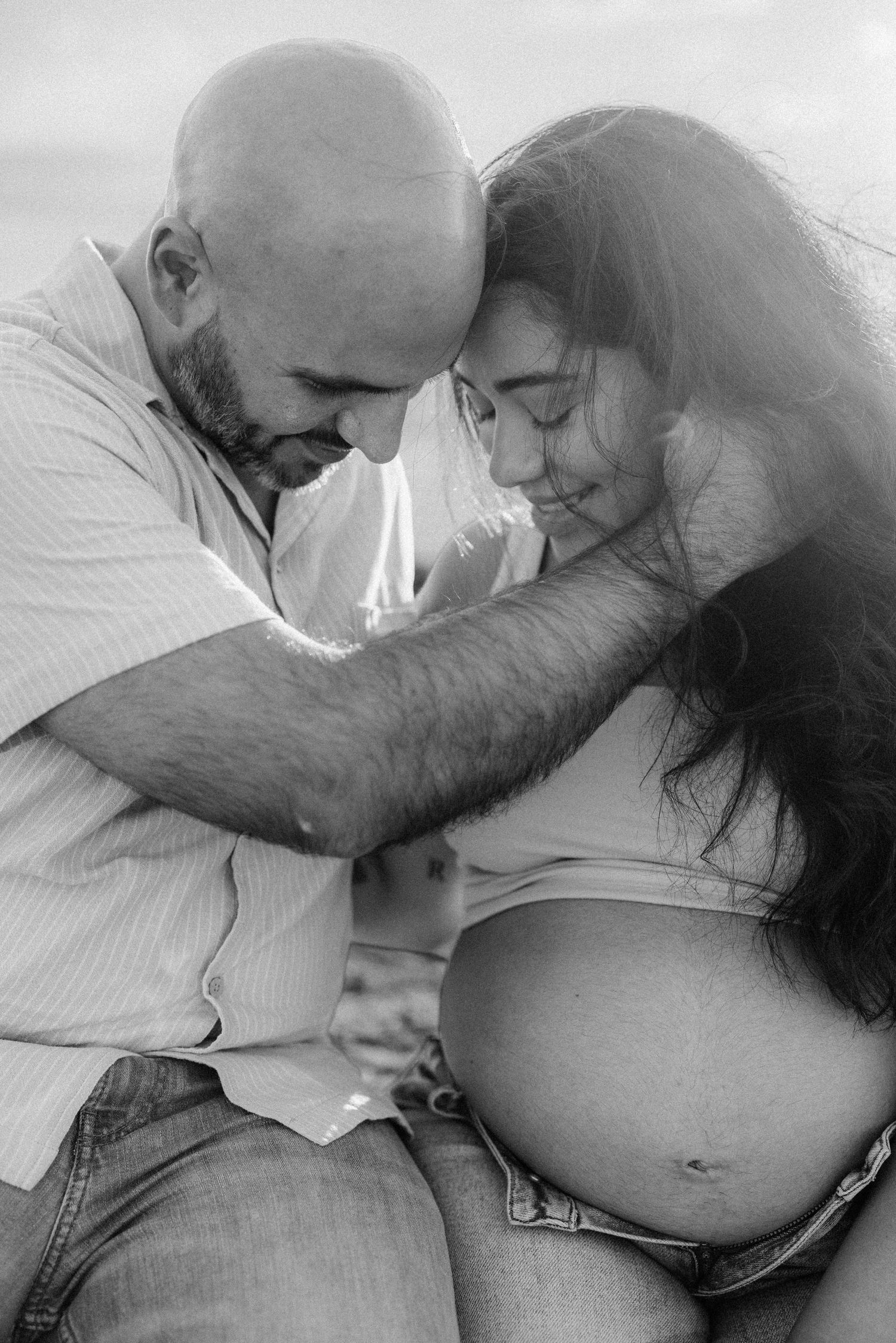 Black and white photo of couple embracing, man leaning on pregnant womans baby bump.