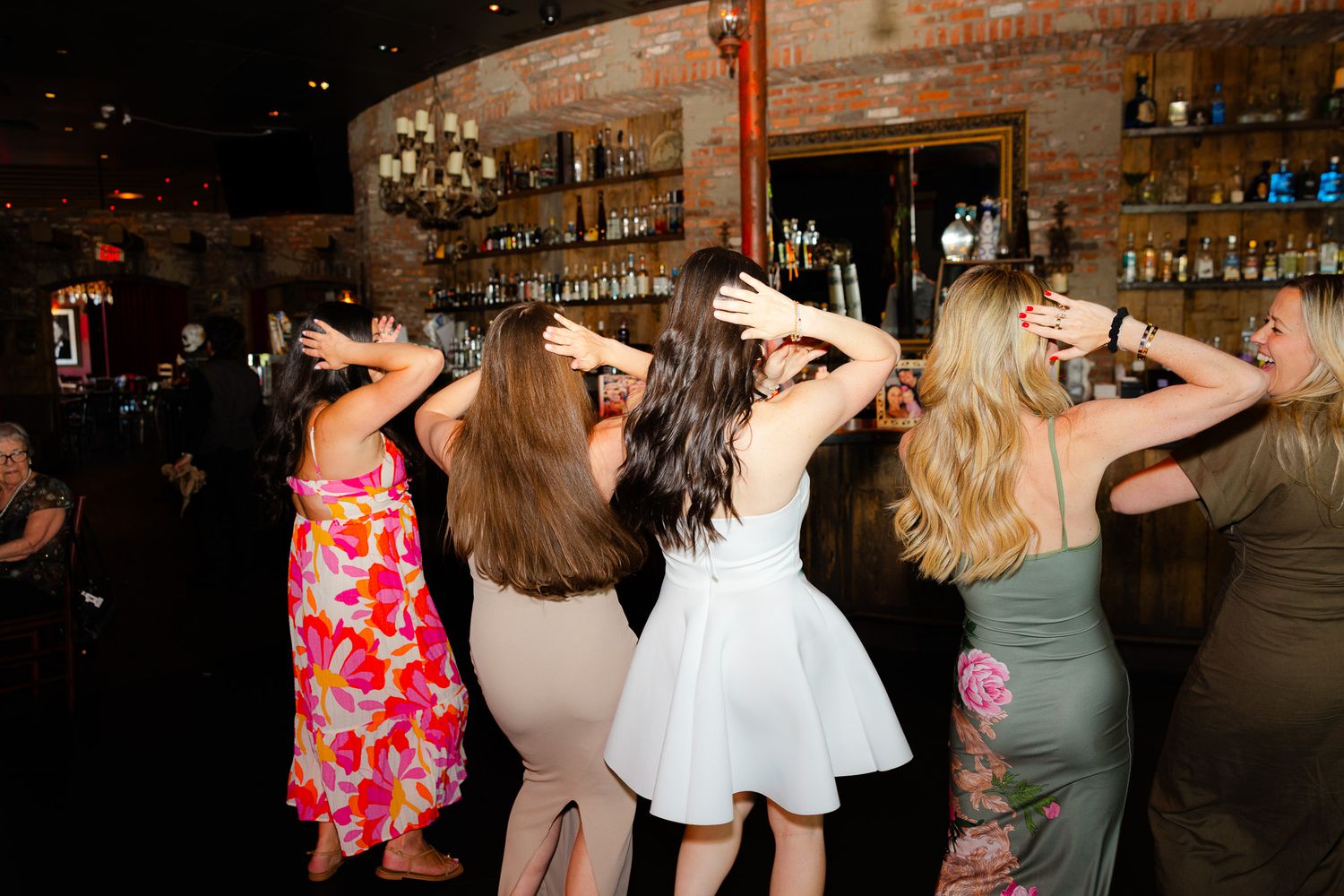 Women dancing at a bar venue, hands raised, wearing colorful dresses at a lively party or wedding reception.