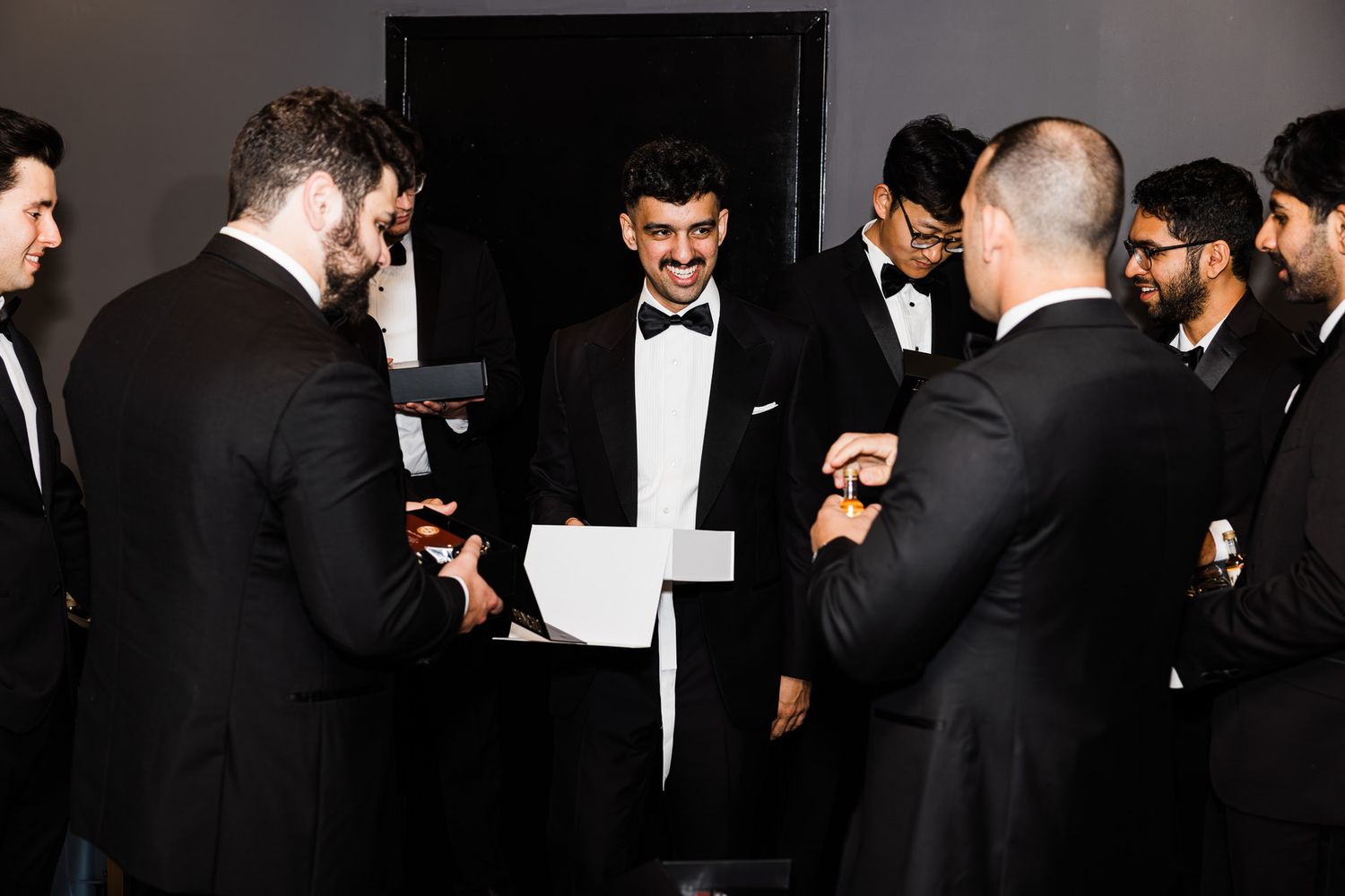 Groomsmen in black tuxedos gather and chat together in a dimly lit room before a wedding ceremony.