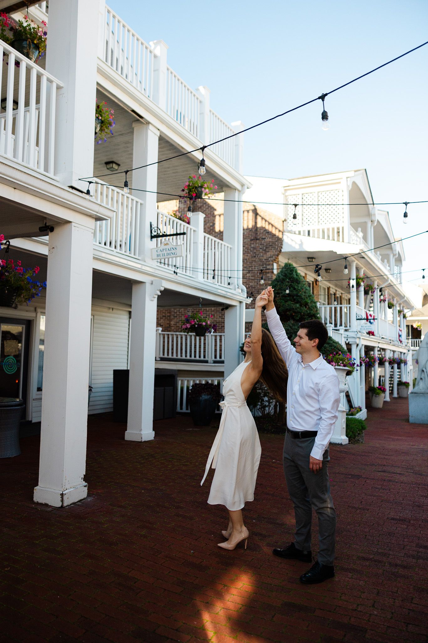 Couple dancing and twirling in a charming brick courtyard with white balconies and string lights at golden hour.