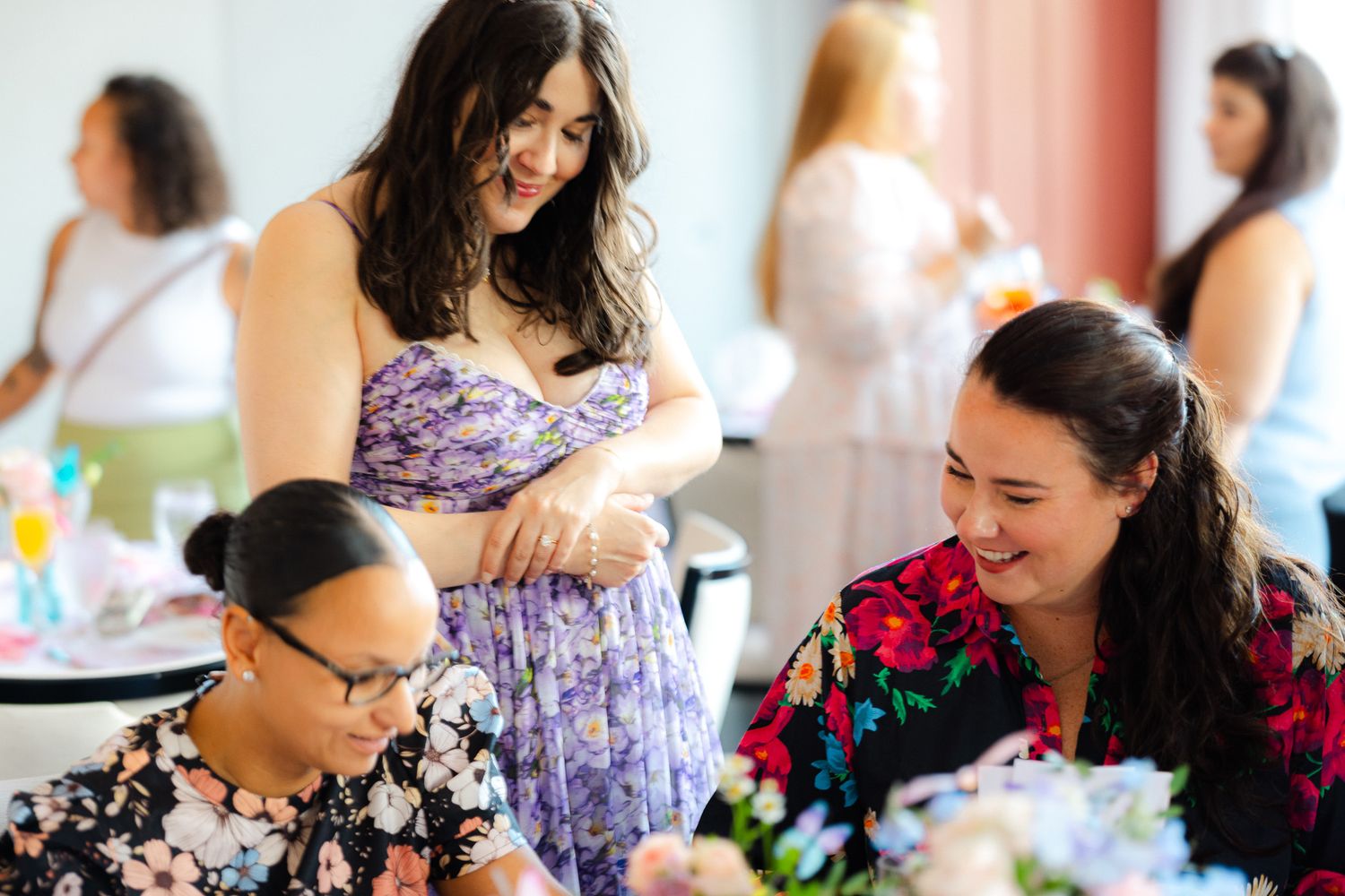 Three women socializing at an elegant event, smiling and conversing among floral arrangements in a bright venue.