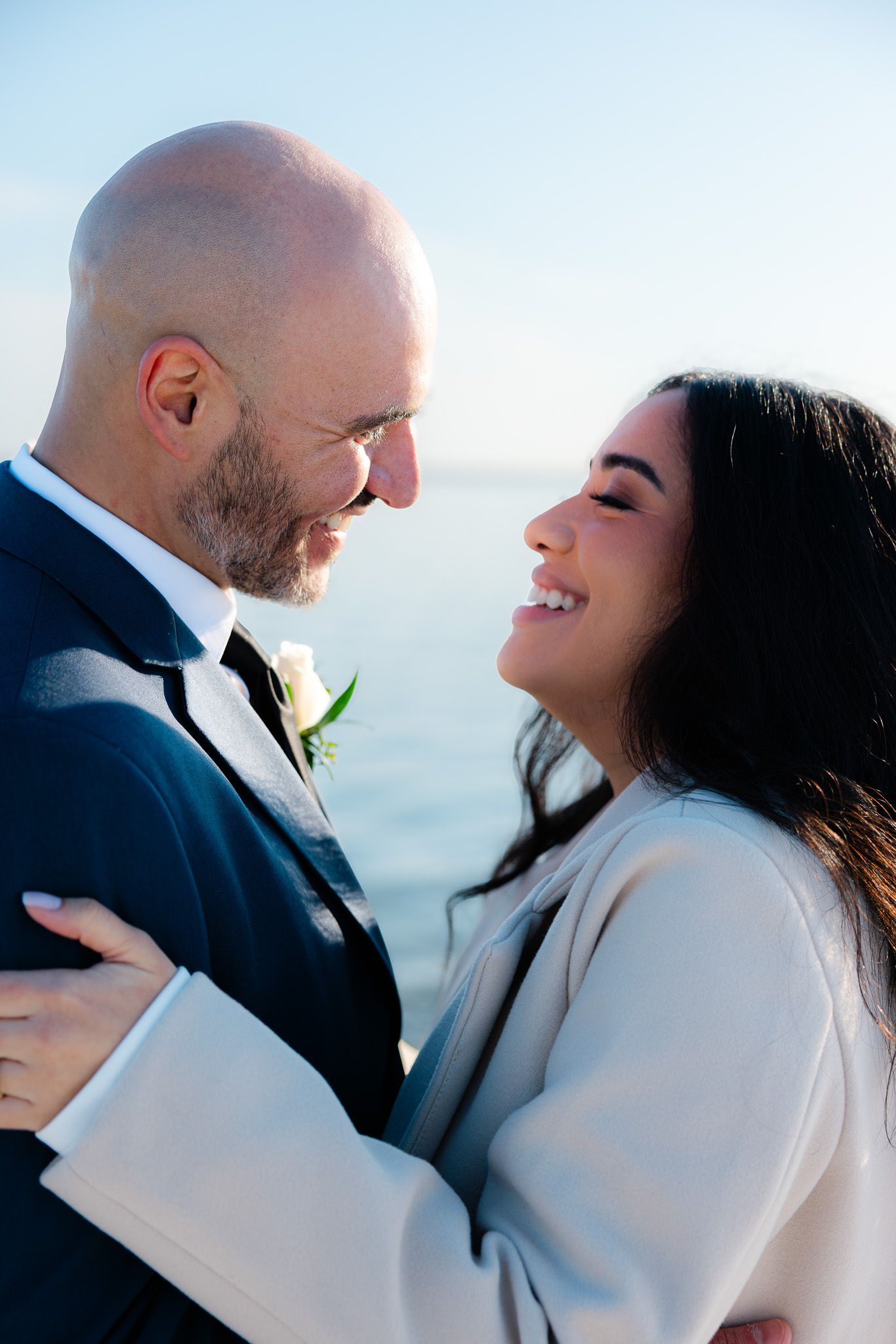 A smiling bride and groom share a romantic moment by the ocean, dressed in elegant wedding attire.