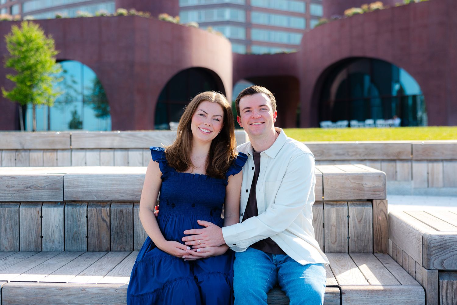 Smiling pregnant couple sitting on wooden steps outdoors near modern brick building on a sunny day.