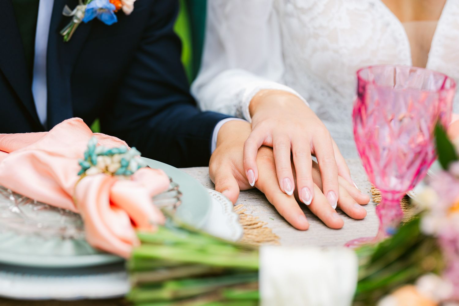 Newlywed couple holding hands at wedding reception table with pink flowers and glassware.