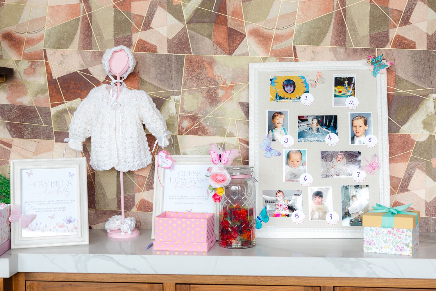 Baby girl party display with pink outfit, photo collage frame, gift boxes, and candy jar on a wooden dresser.