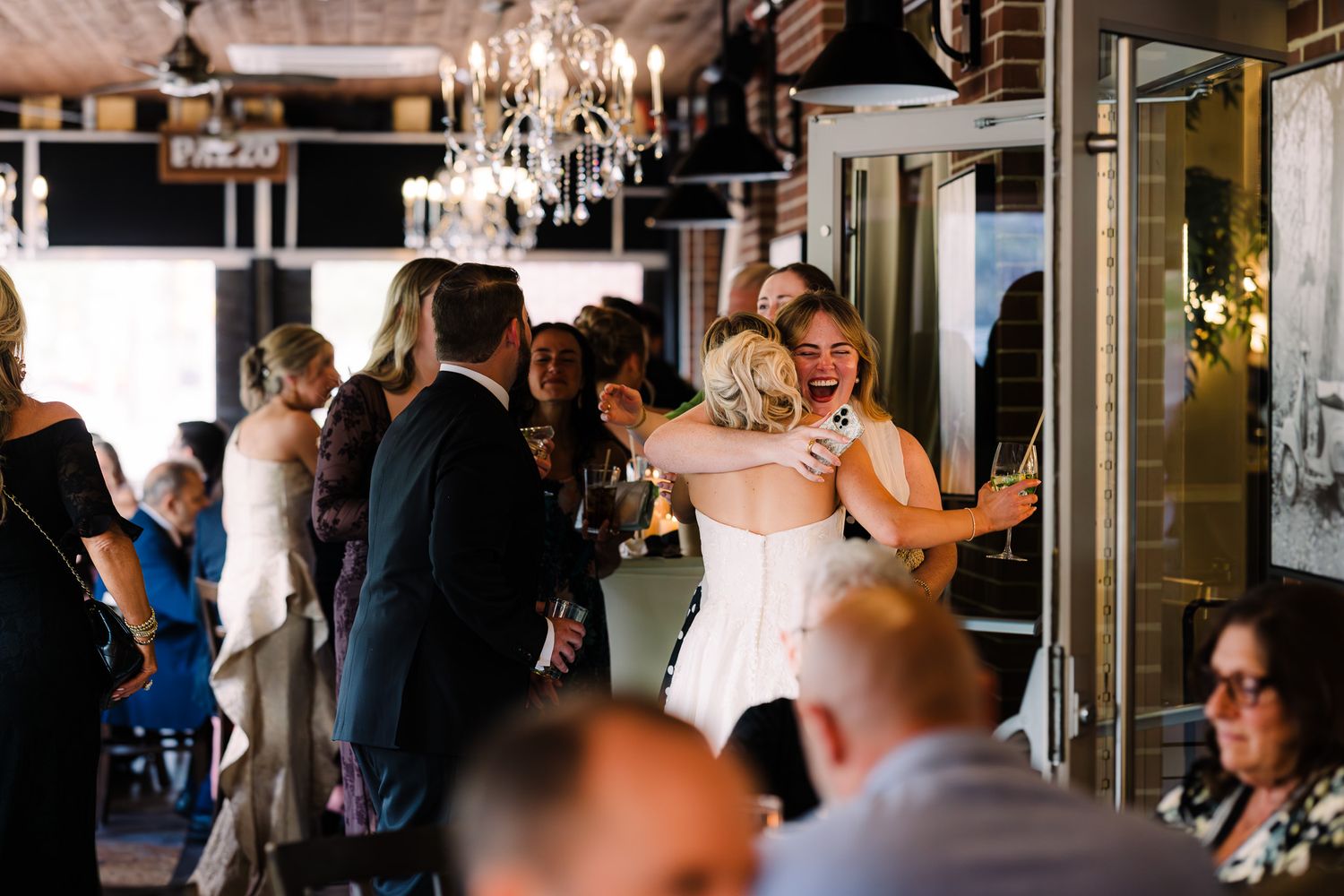 Guests mingling and hugging at an elegant wedding reception with chandeliers and warm lighting.