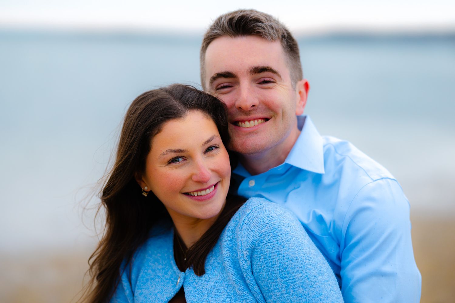 Happy couple smiling at camera wearing matching blue shirts on beach with ocean in background.