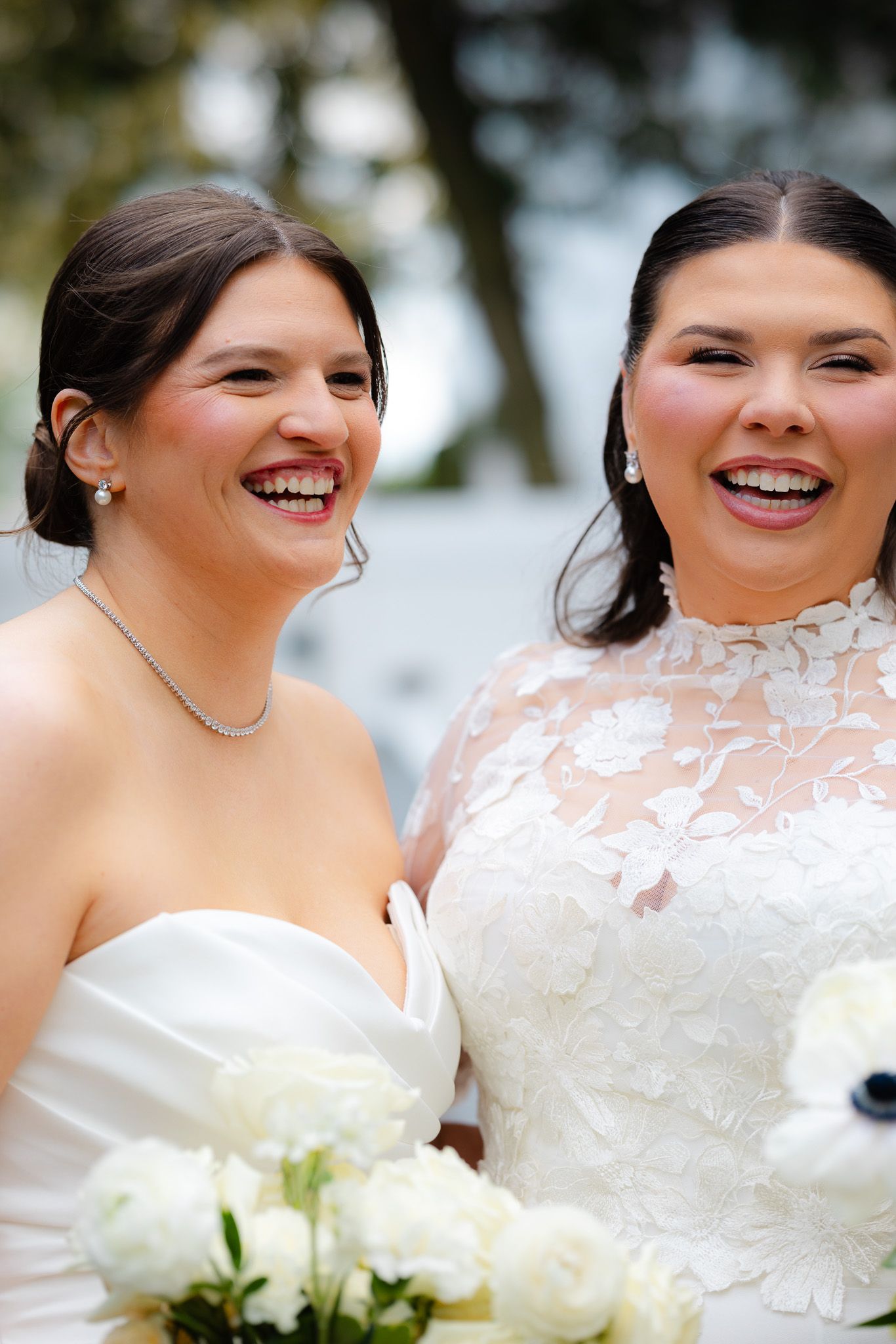 Two brides in white wedding gowns laughing joyfully, one holding white floral bouquet, celebrating their wedding day.