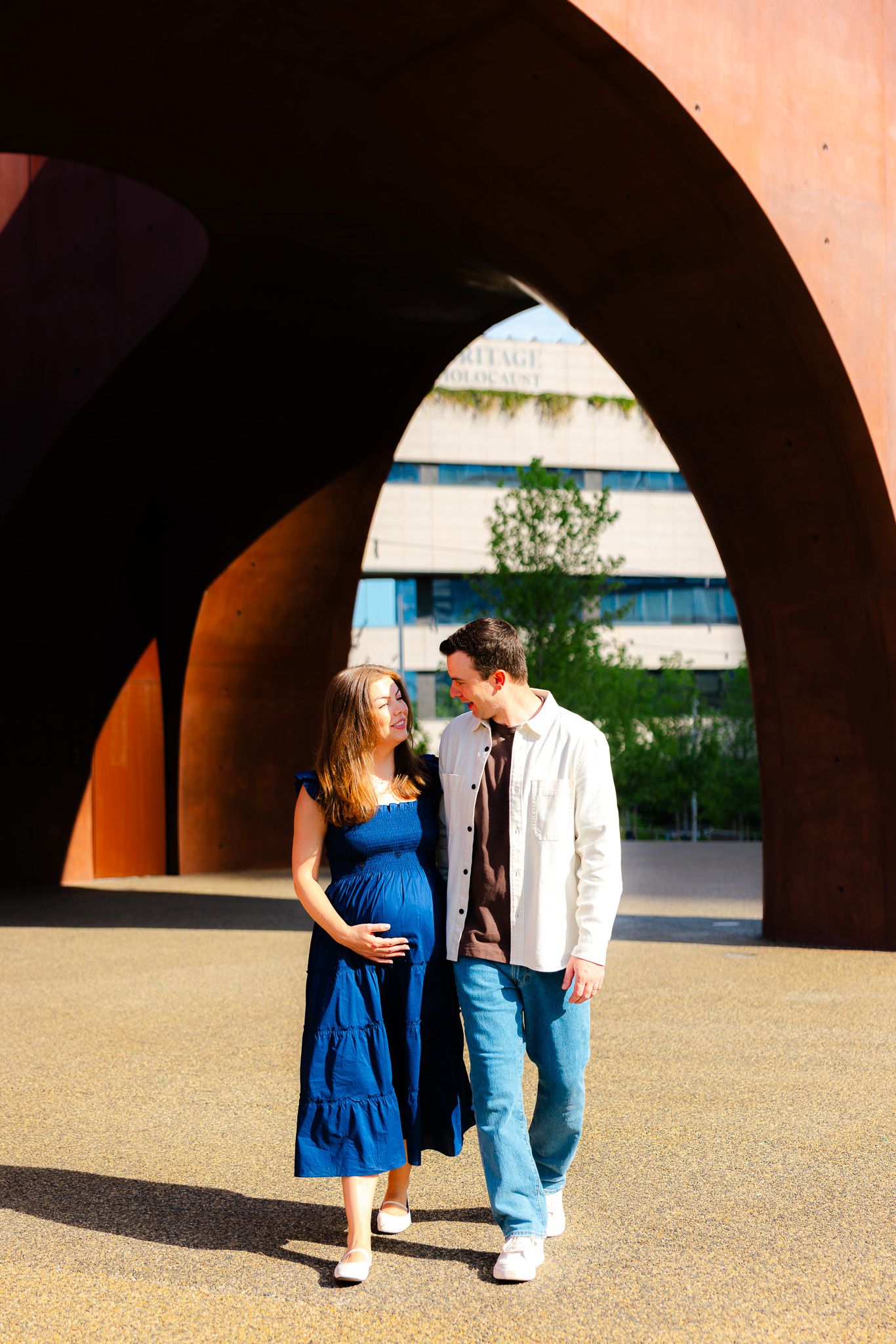 Pregnant woman in blue dress and man in white jacket stand together under rustic arches in urban setting.