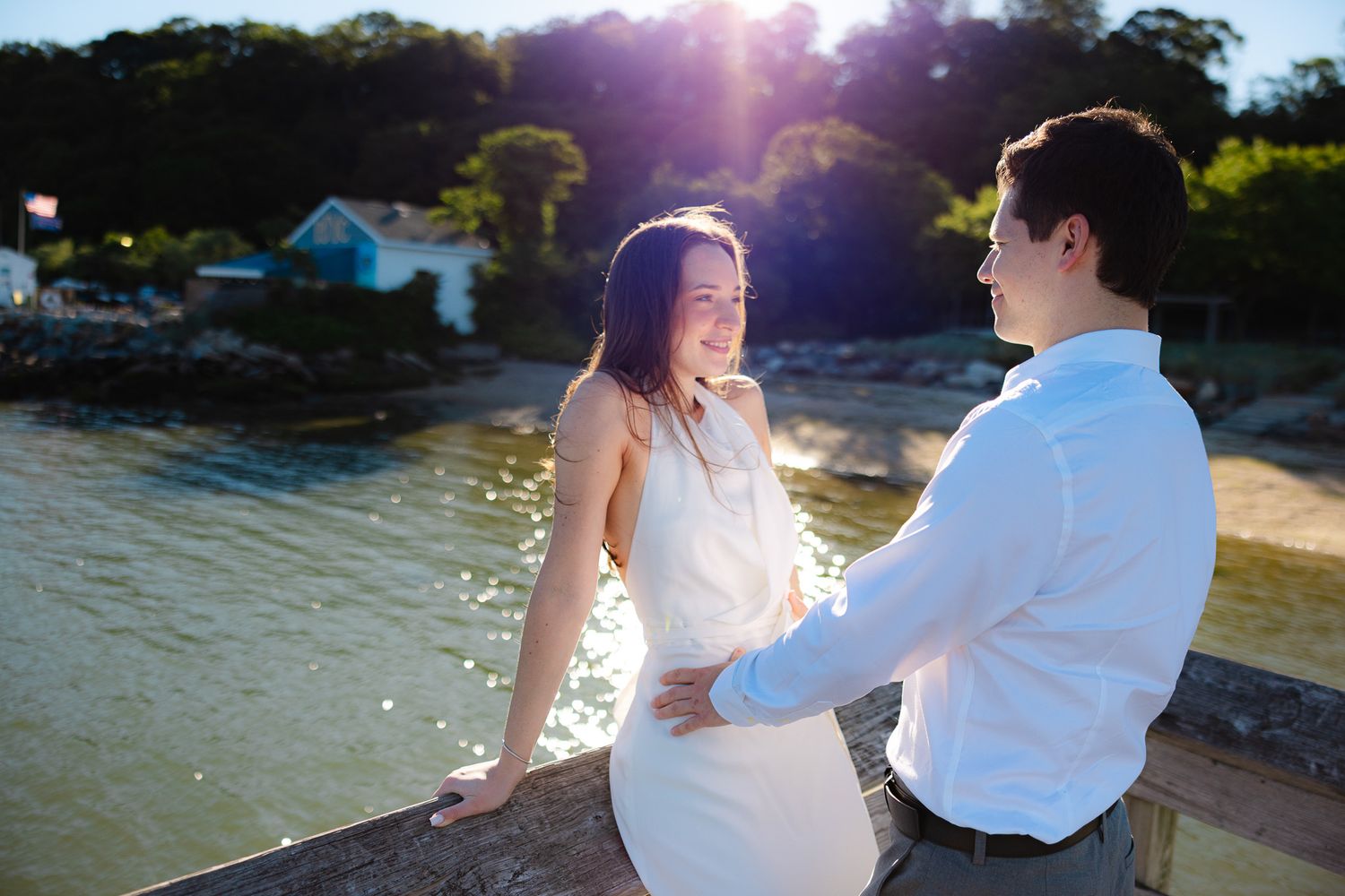 Couple sharing a romantic moment by the waterfront at golden hour, woman in white dress smiling.