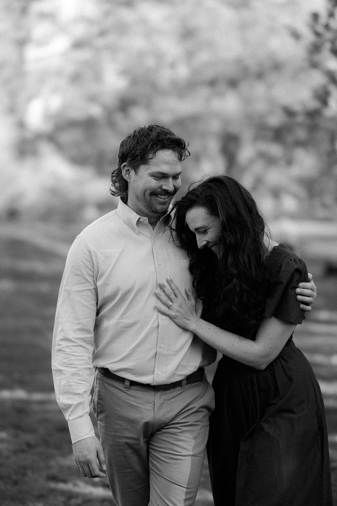 Black and white photo of a smiling couple walking together in a park, woman leaning on mans shoulder.