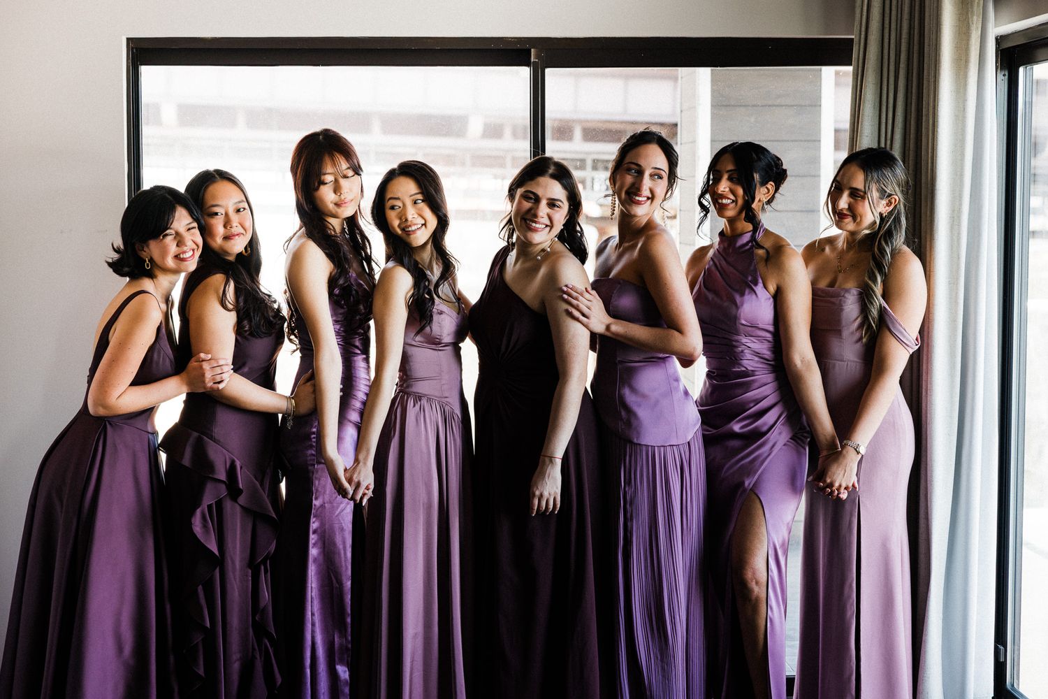 Seven bridesmaids pose together in matching purple gowns in front of a large window.