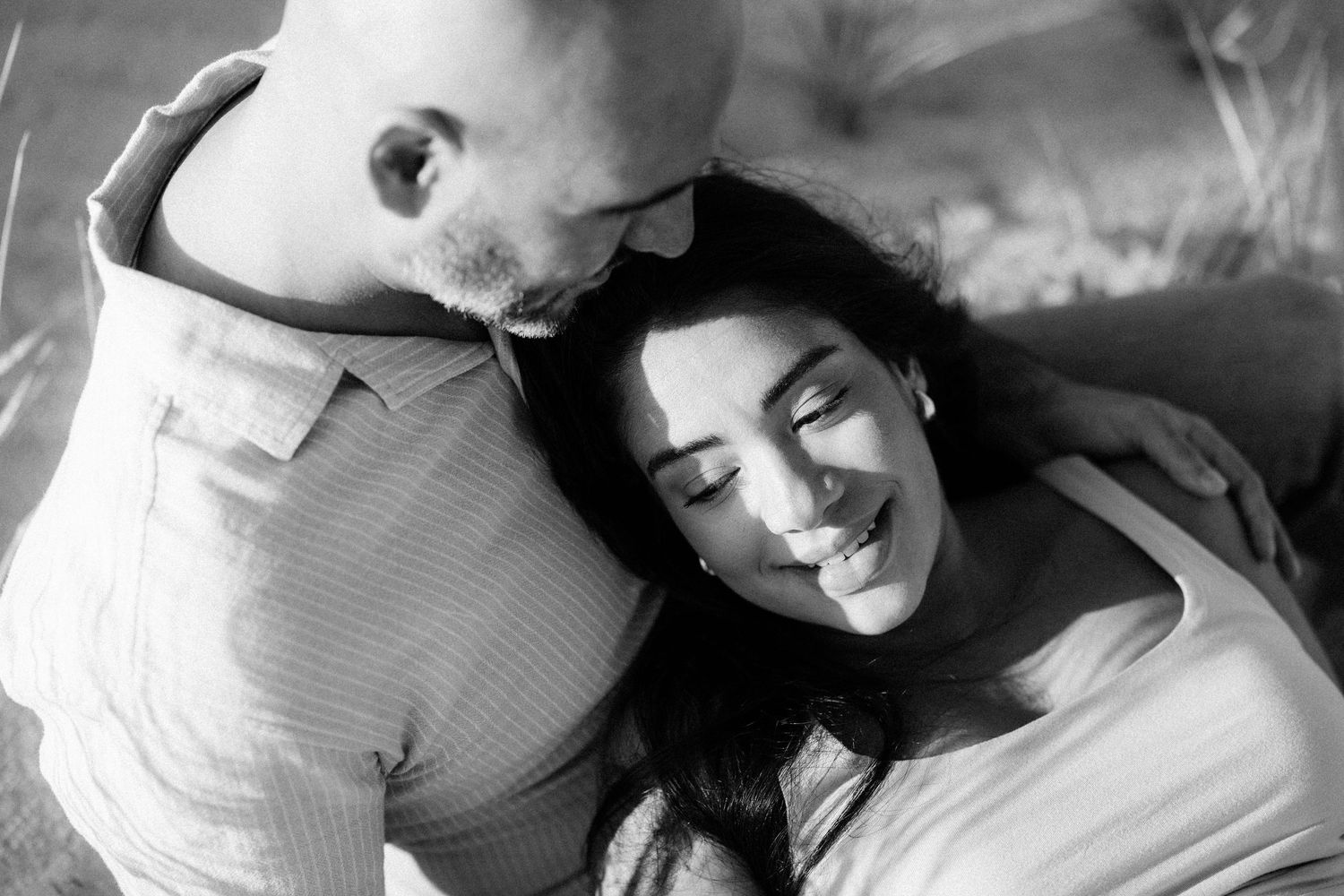 Black and white photo of a couple embracing outdoors, man kissing woman's head as she smiles.