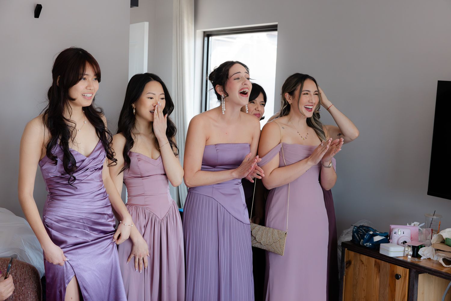 Four bridesmaids in purple satin dresses laughing and reacting joyfully during wedding preparations indoors.