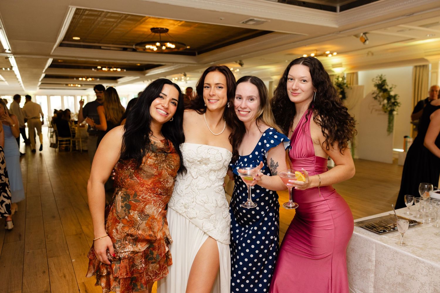 Four women in elegant dresses smile together at a wedding reception in a decorated venue.