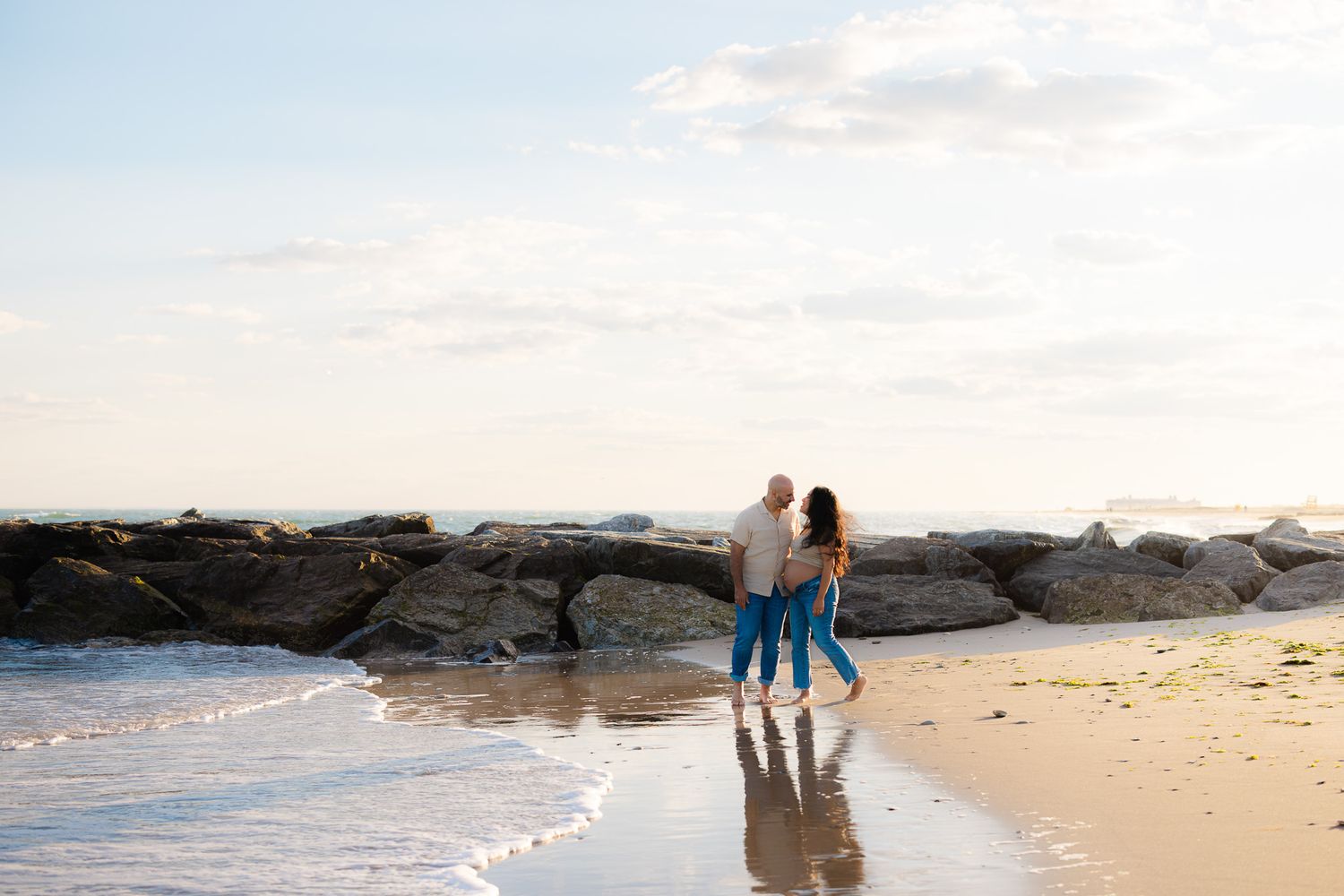 Couple standing together on a sandy beach near rocky jetty with gentle waves and soft golden sky.