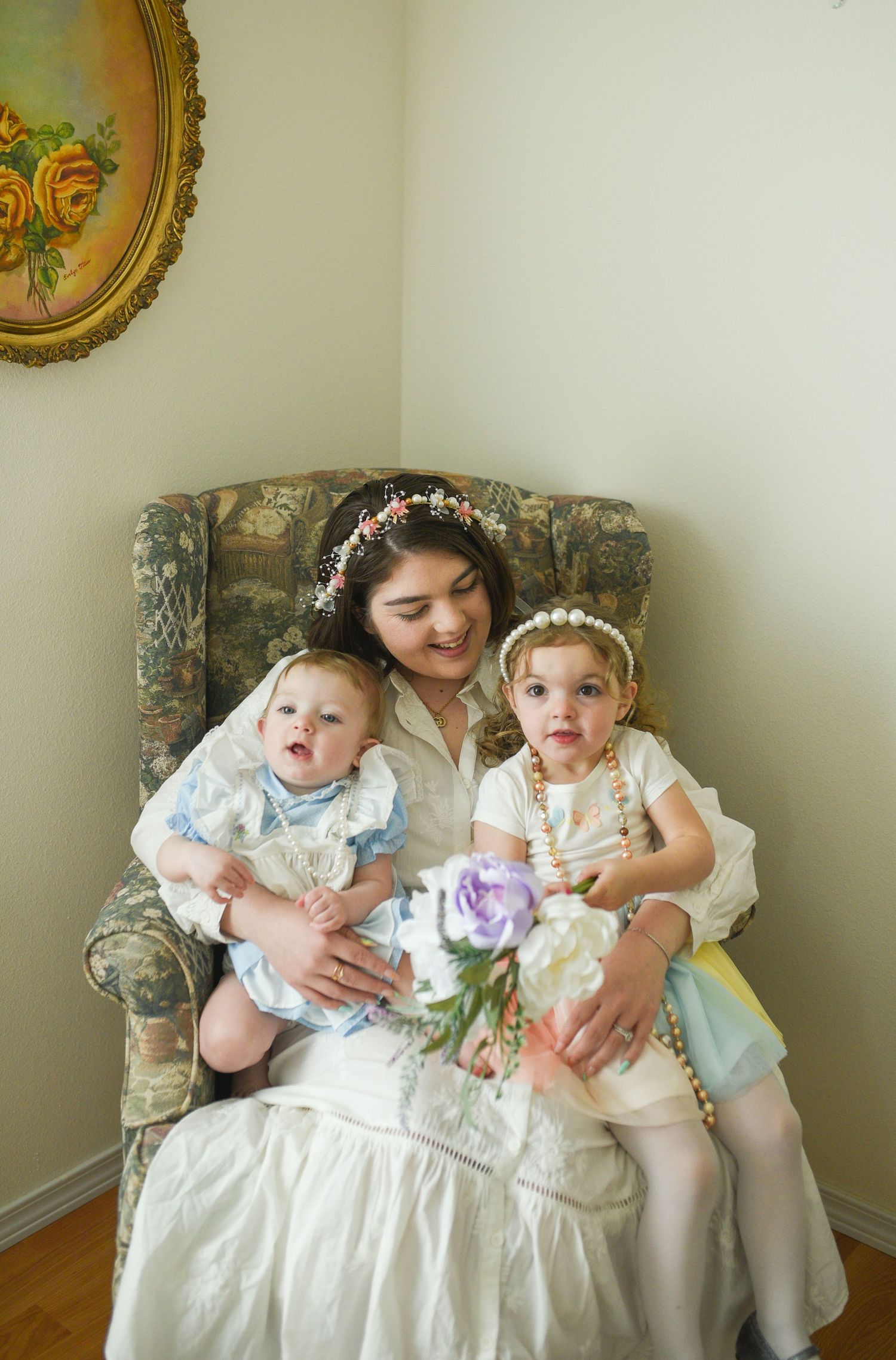 A mother wearing a floral crown sits in an armchair holding two young daughters in vintage dresses.