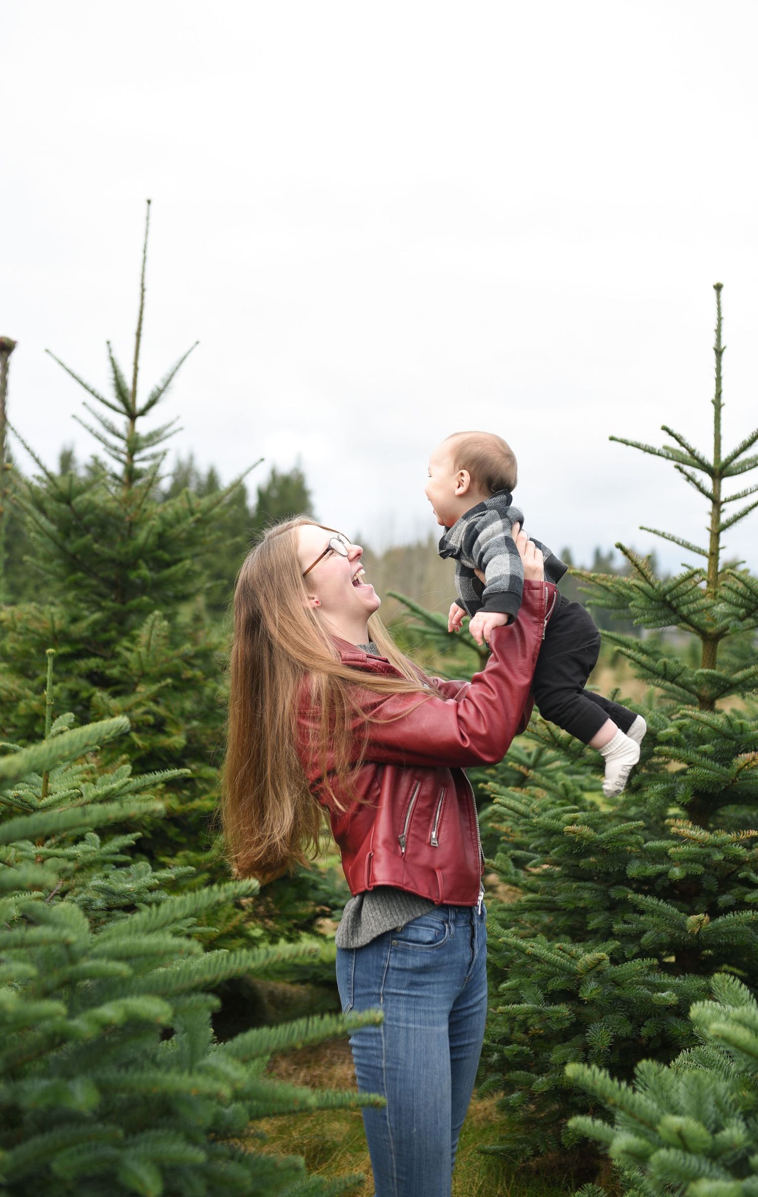 A woman in a red leather jacket lifts a smiling baby into the air at a Christmas tree farm.