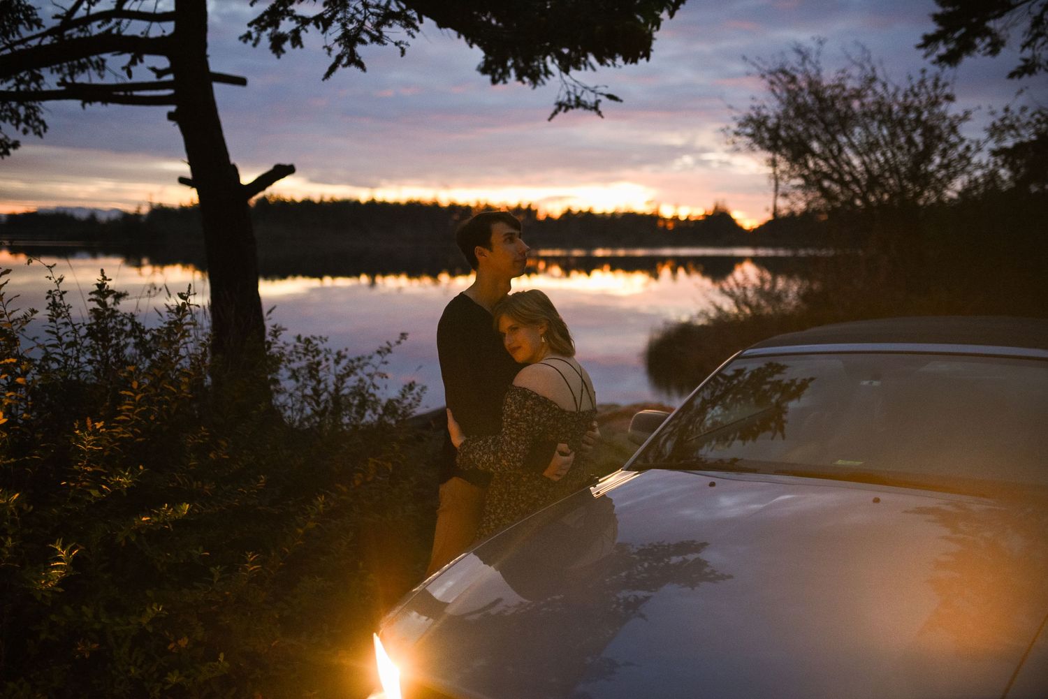 Couple embracing by a car near a lake at sunset with colorful sky reflected on water.