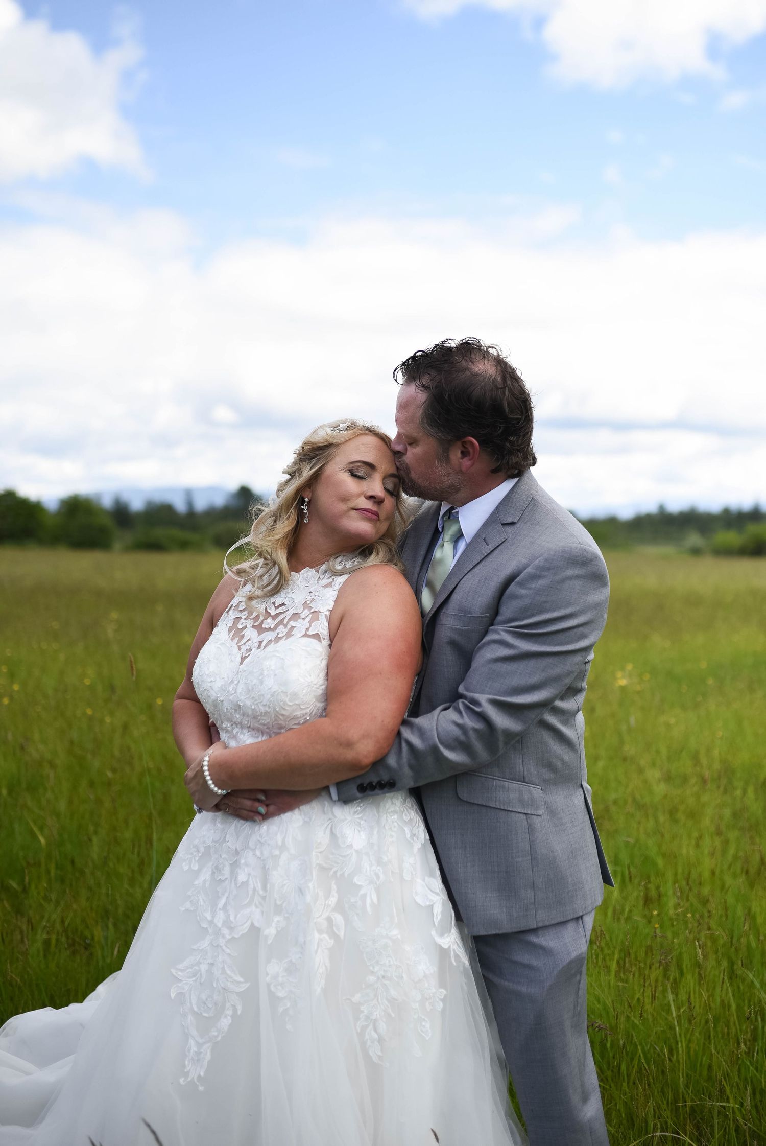 Bride and groom share a romantic embrace in a lush green meadow, groom kissing bride's cheek in wedding attire.