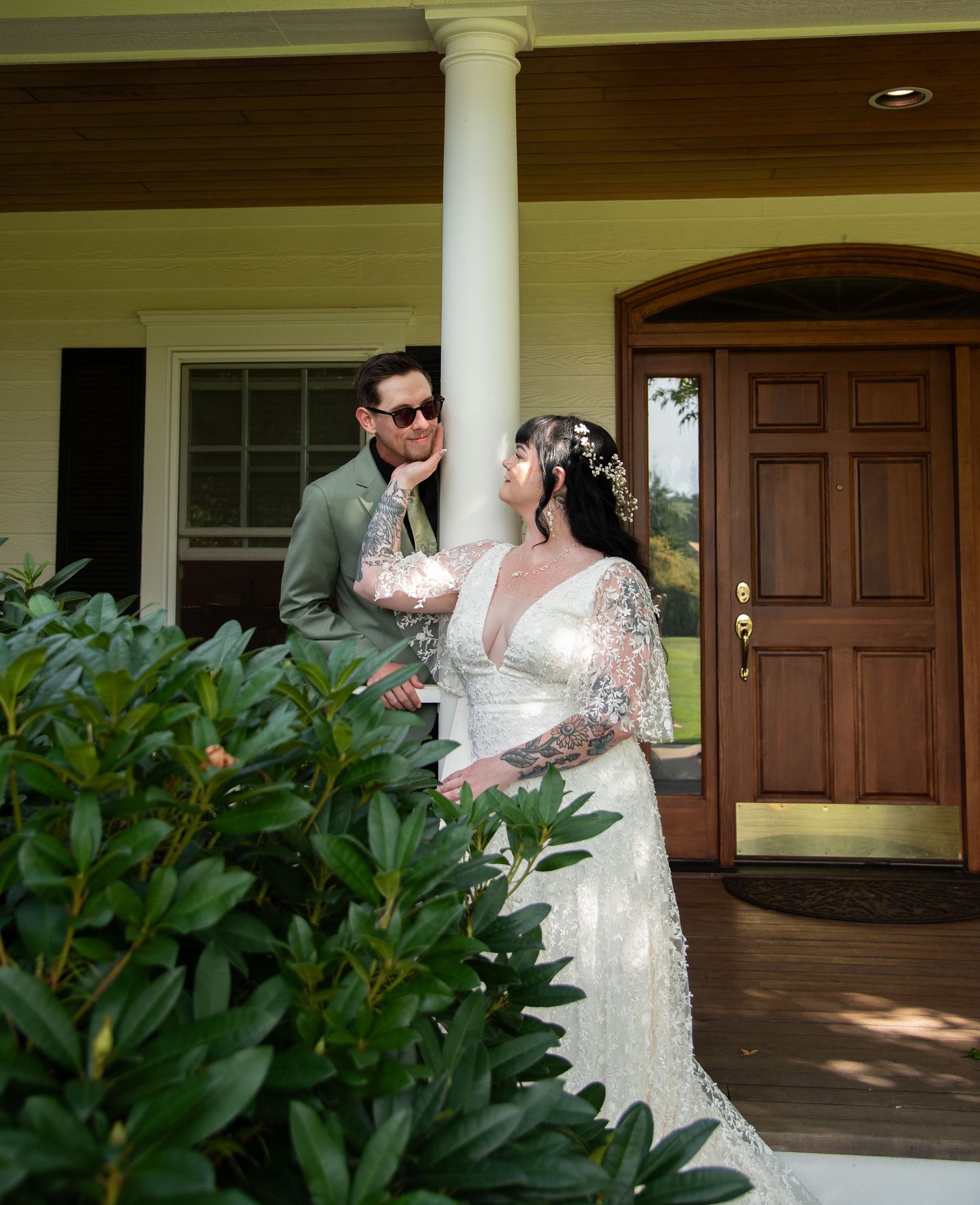 Bride and groom posing on a porch, woman in lace wedding gown with floral hair accessories and tattooed arms.
