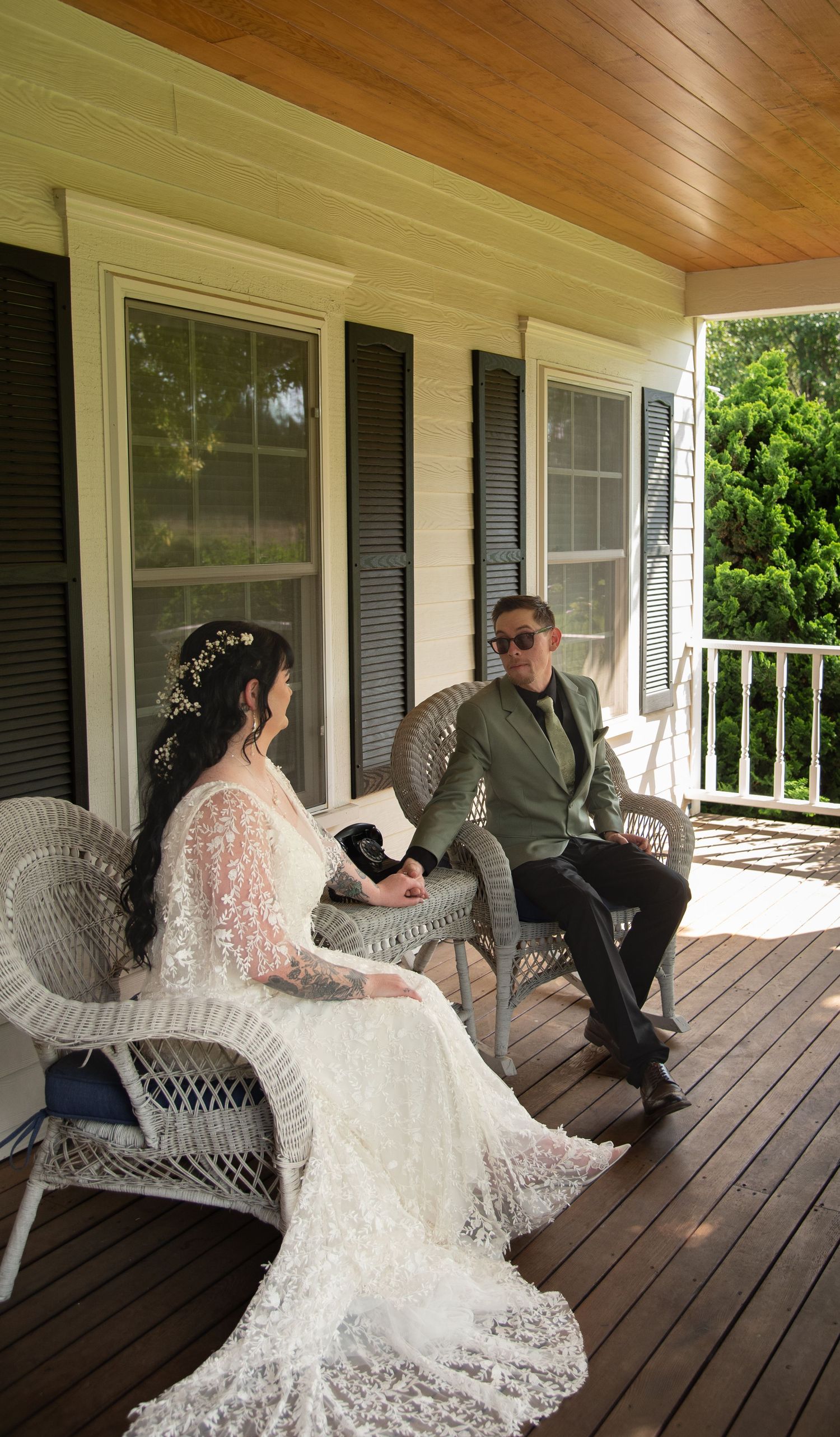 Bride and groom in wedding attire sit together on wicker chairs on a wooden porch.