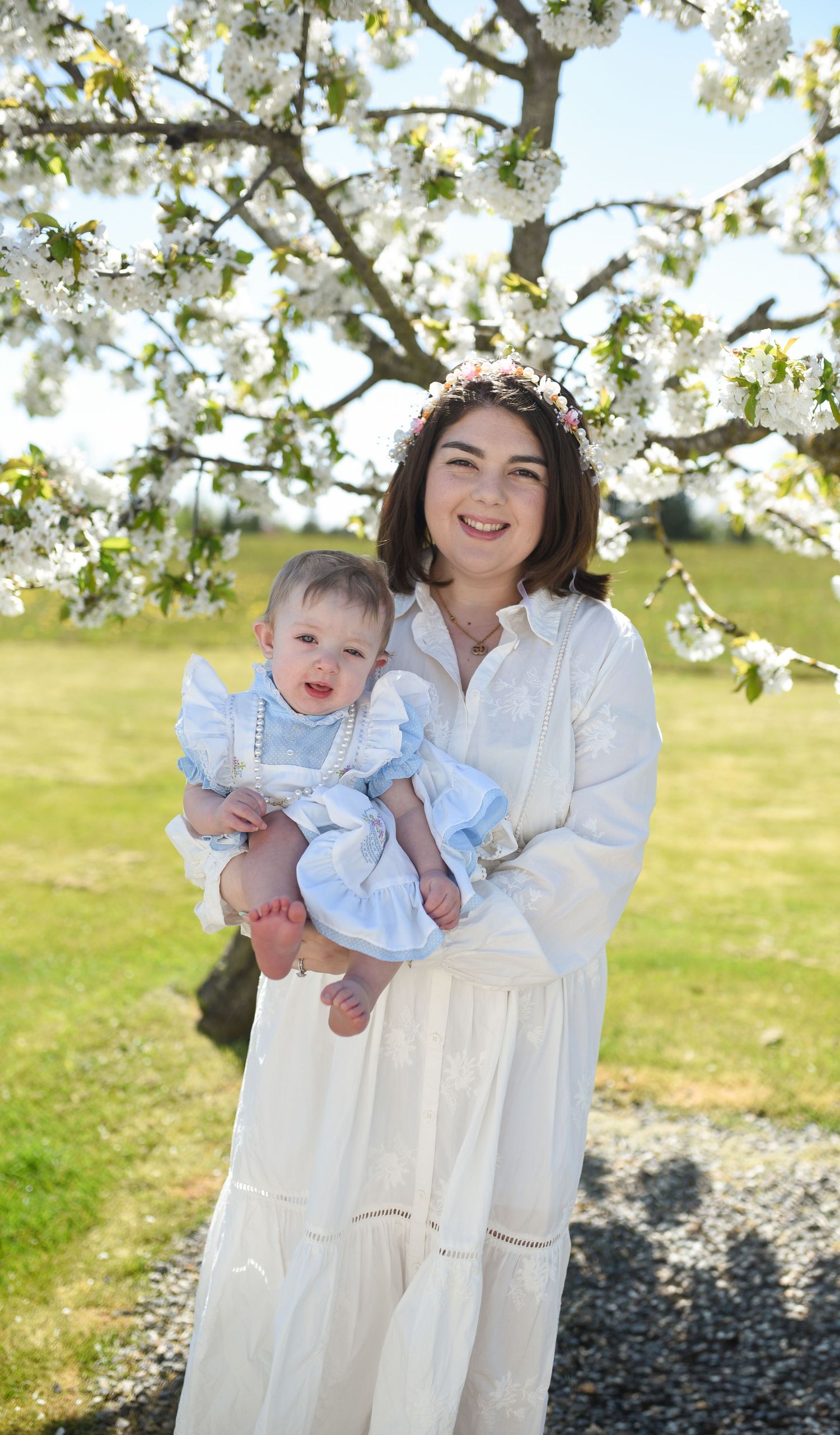 Smiling woman in white dress holding a baby under blooming cherry blossom tree outdoors.