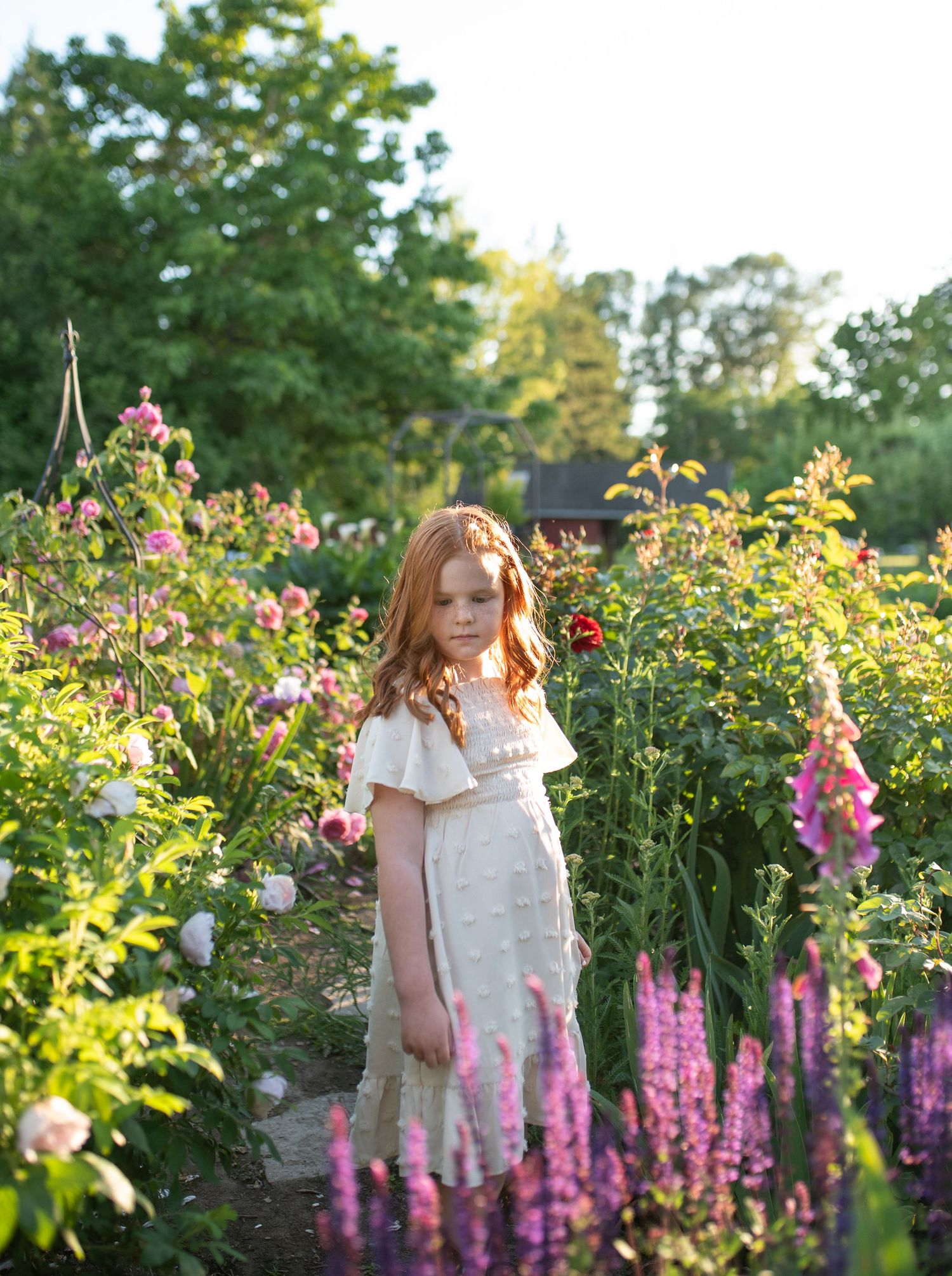 Red-haired girl in white dress standing among colorful garden flowers at sunset.