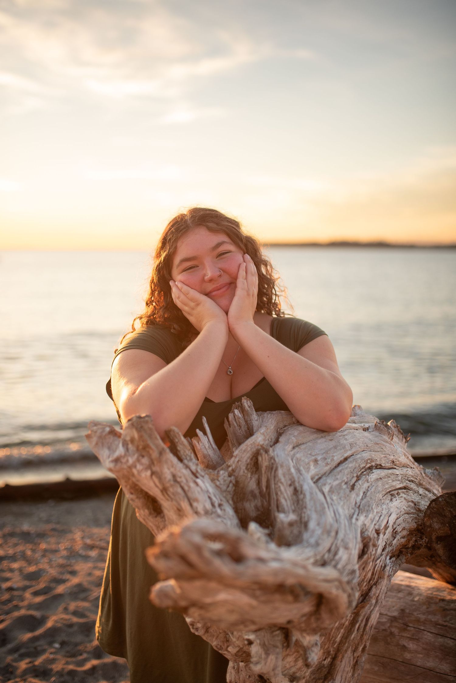 Smiling young woman resting face in hands on driftwood at beach during warm sunset.
