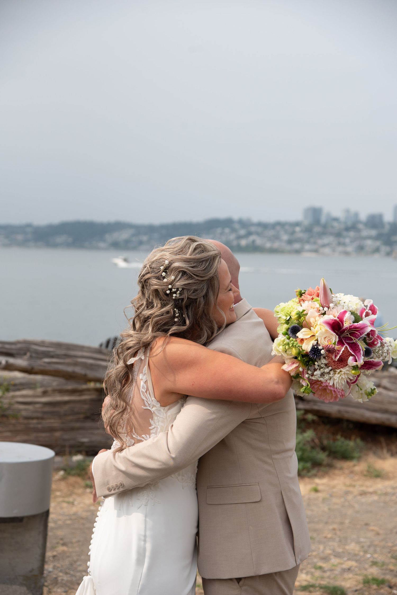 Bride and groom share an emotional first look hug outdoors by the waterfront, bride holding colorful bouquet.