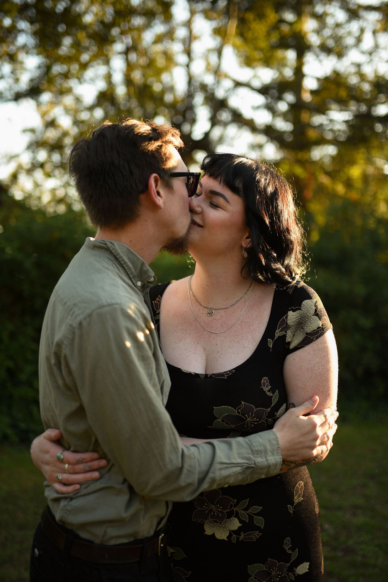 Couple sharing a romantic kiss outdoors in golden sunset light surrounded by green trees.