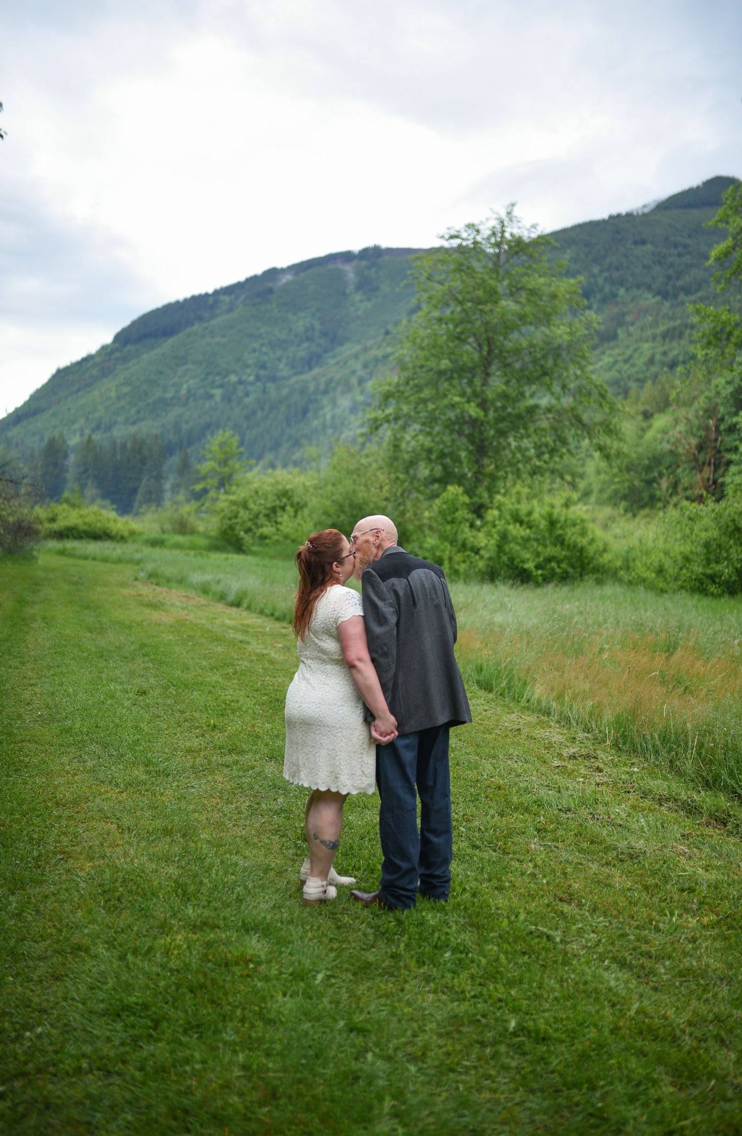 Couple sharing a kiss in white dress and gray suit on green lawn with mountain backdrop.