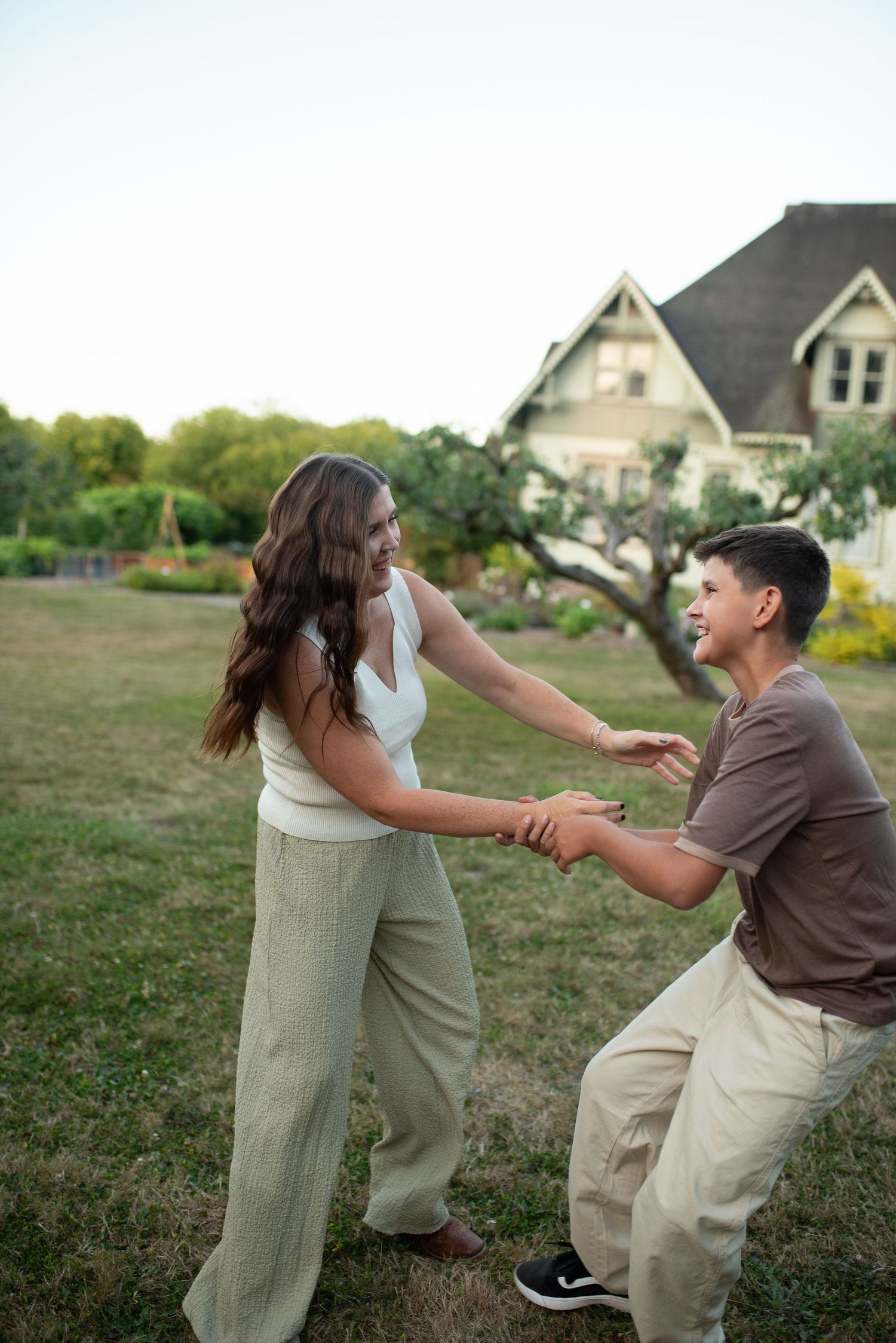 A mother and teenage son playfully hold hands and dance together on a green lawn near a historic home.
