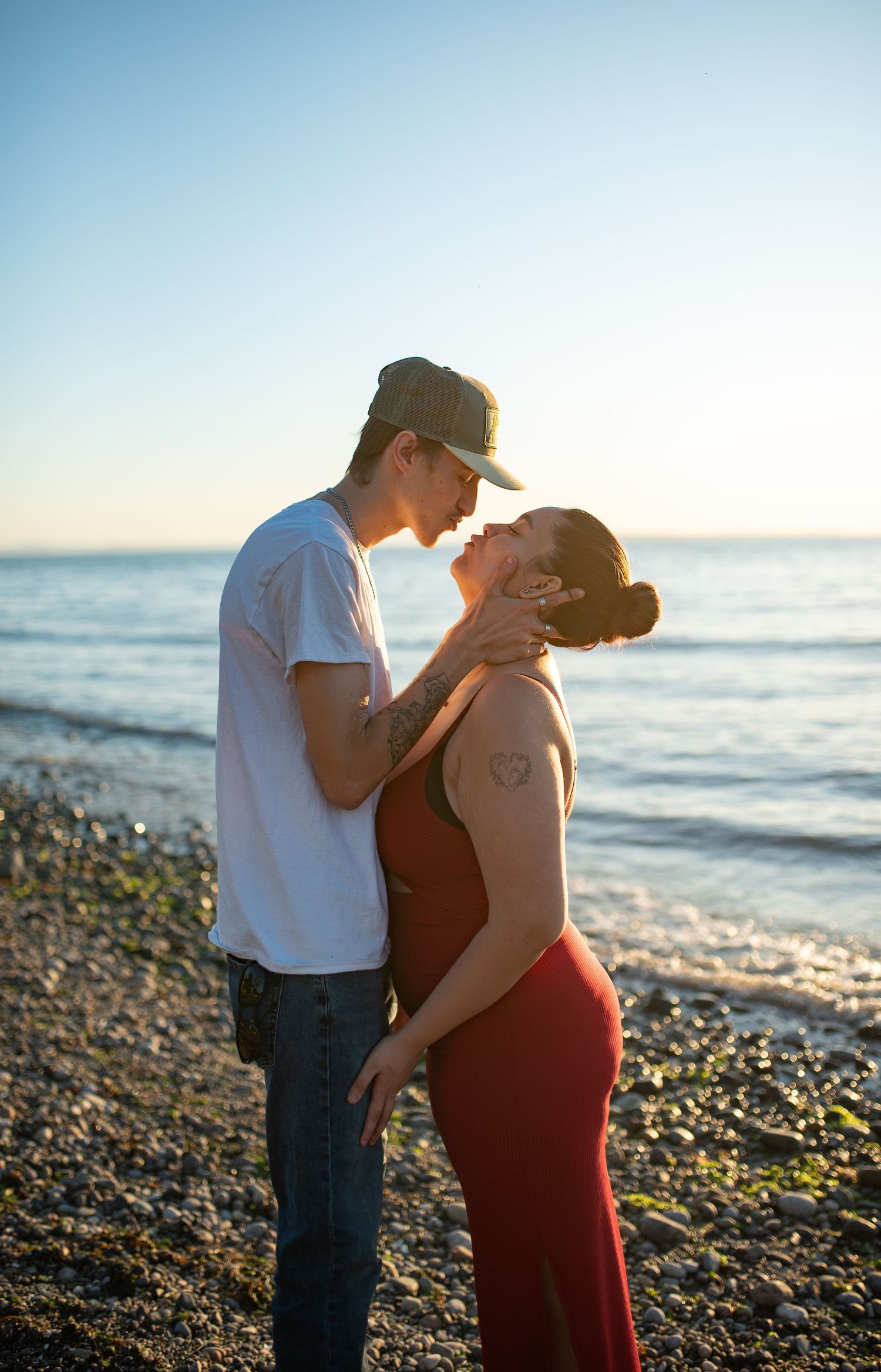 Couple in tender moment on rocky beach at sunset, man gently holding woman's face near the ocean.