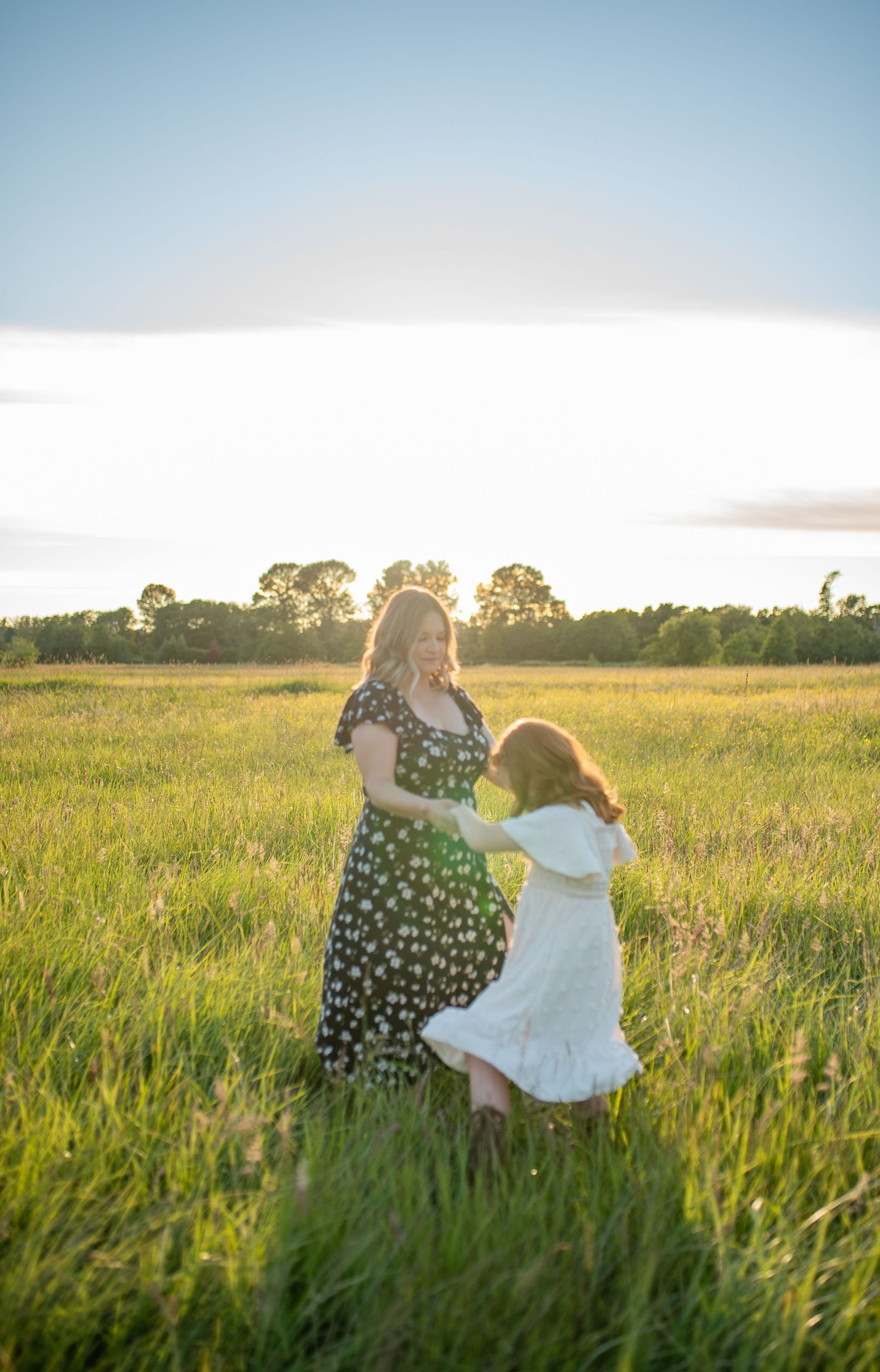 Mother and red-haired daughter in white dress dance together in a glowing sunset meadow.