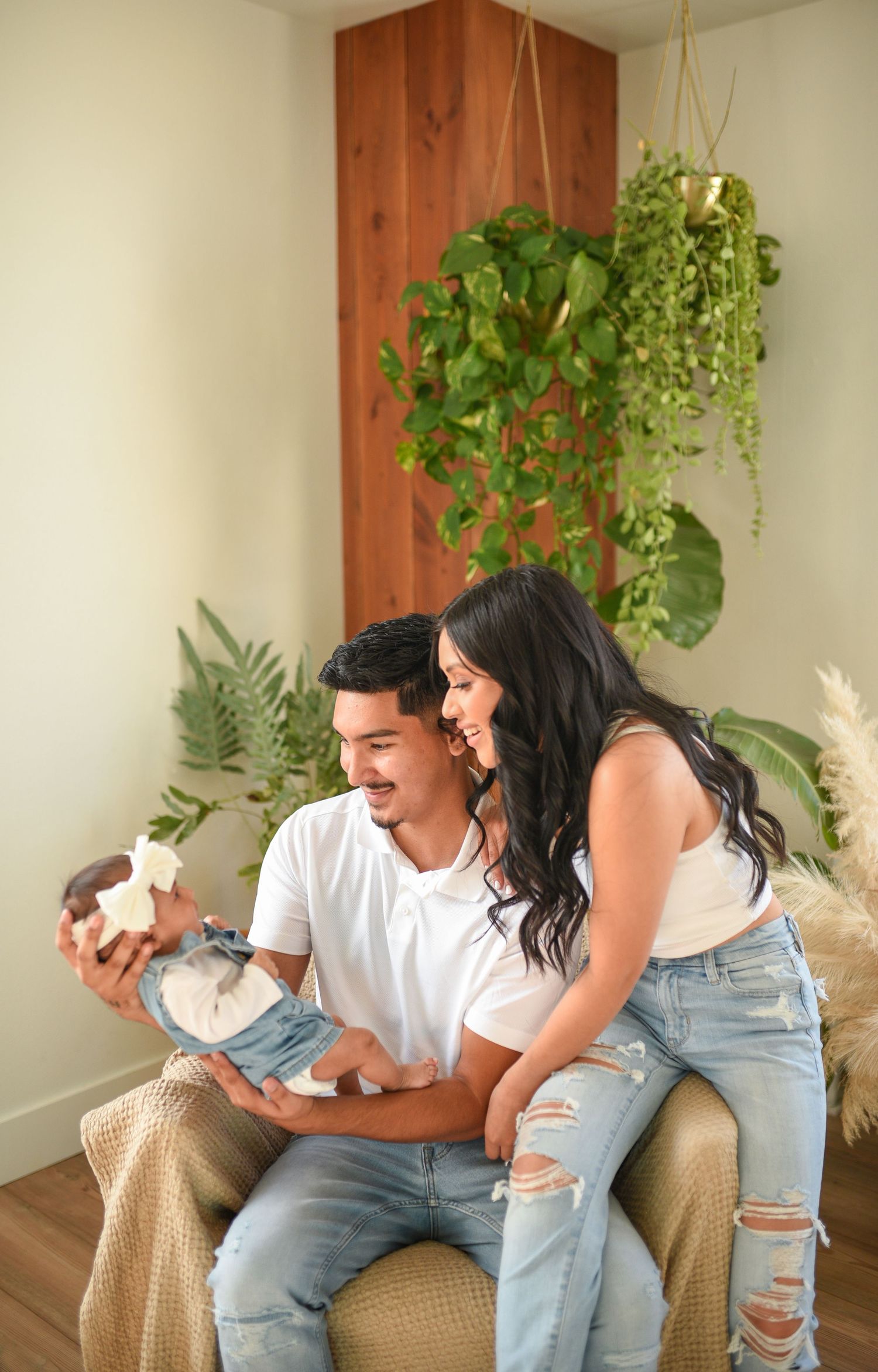Happy couple sitting in a cozy corner with hanging plants, lovingly admiring their newborn baby wrapped in a bow.