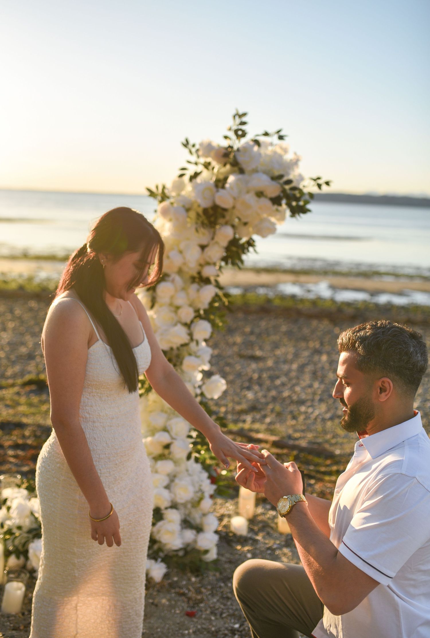 Man proposes to woman on beach at sunset with white floral arch backdrop.