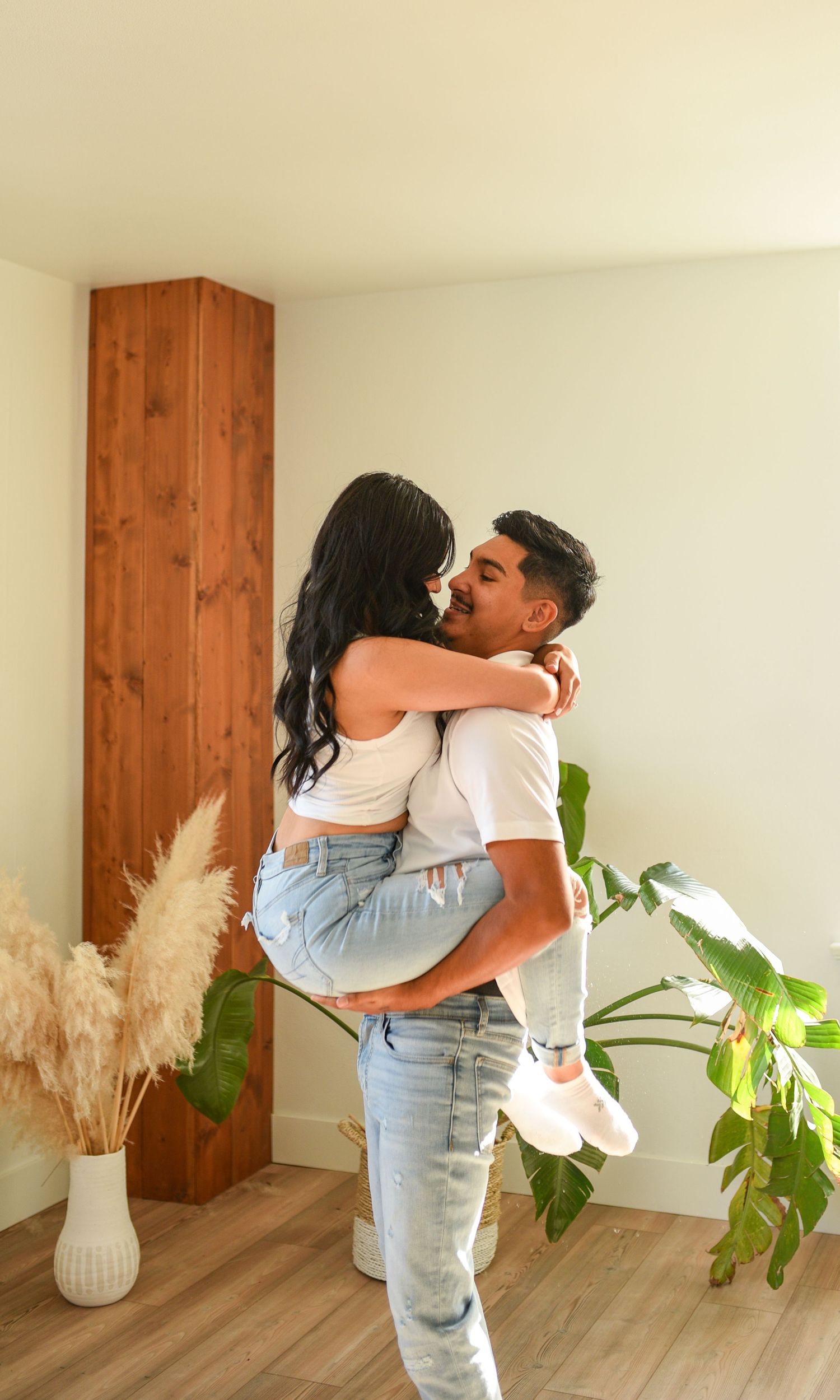 Couple embracing as man lifts woman in cozy room with wood accent wall and tropical plants.