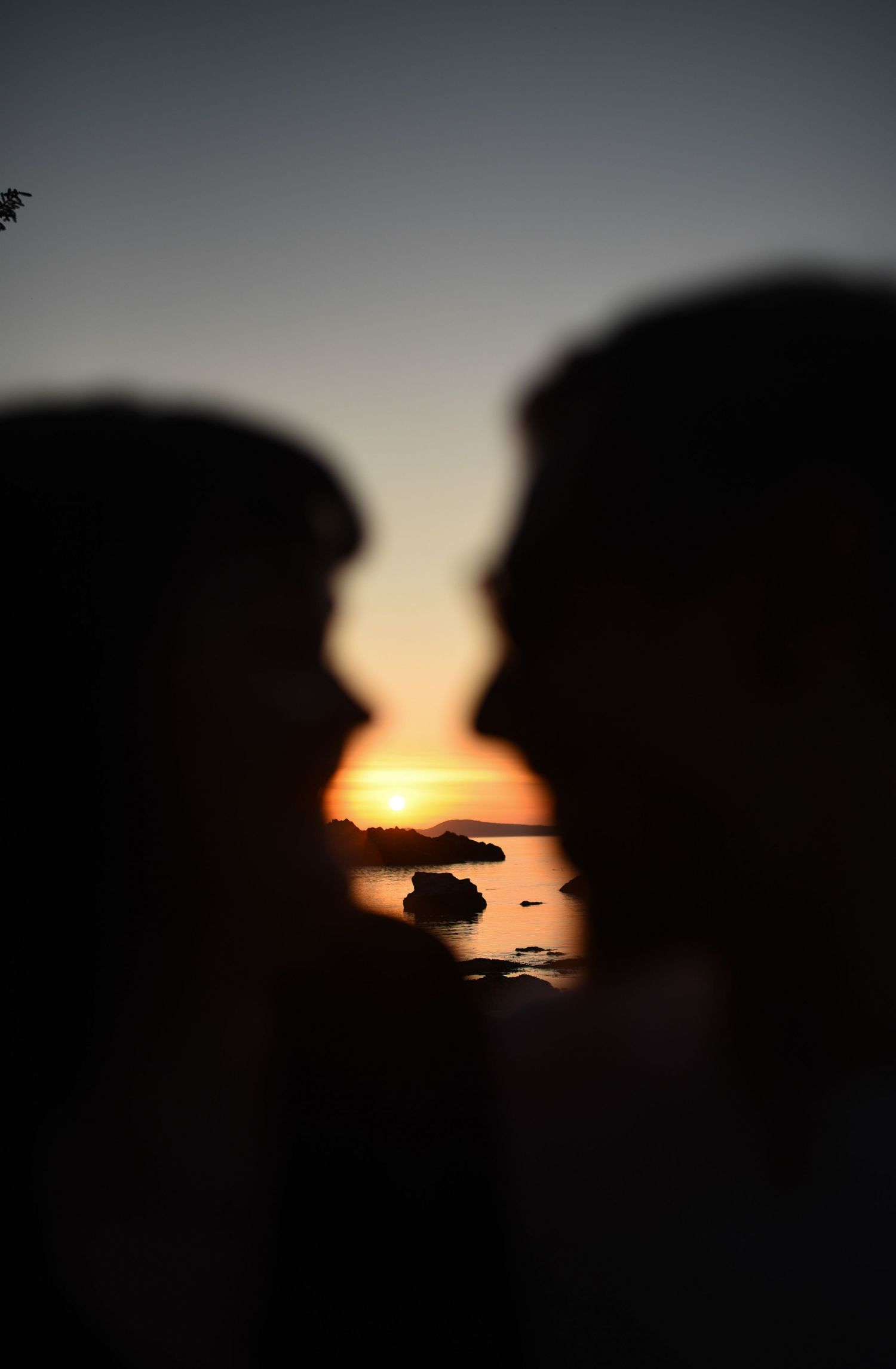 Romantic silhouette of couple about to kiss framing a vivid orange sunset over rocky coast.