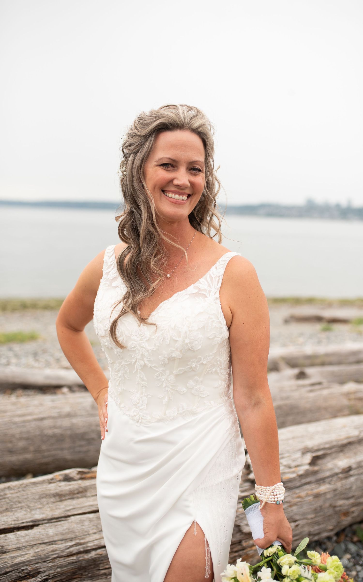 Smiling bride in white lace wedding gown holds bouquet while posing by driftwood on a beach.