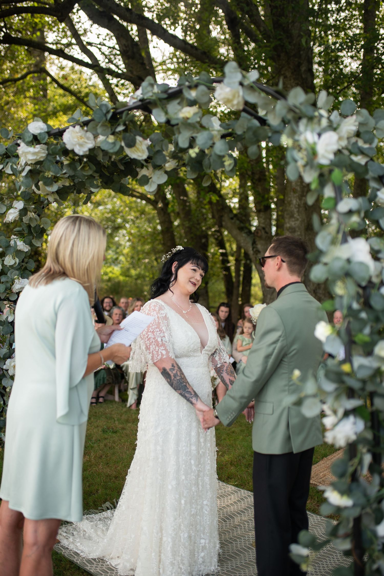 Bride and groom exchanging vows under floral arch with white roses and eucalyptus at outdoor wedding.