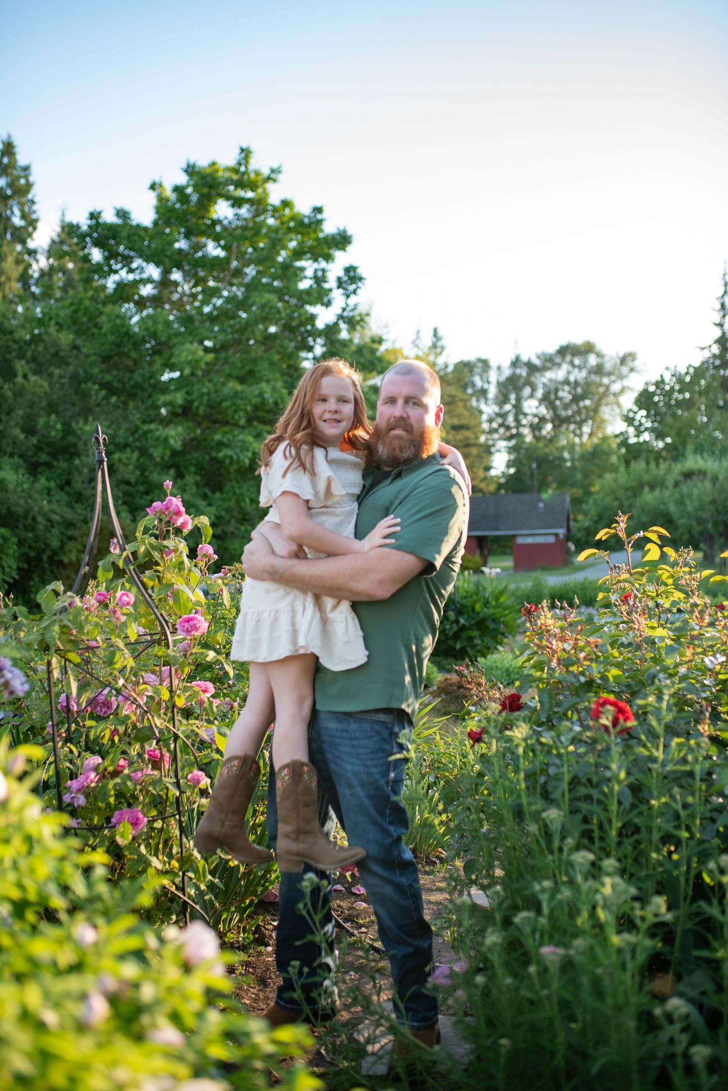 Bearded father holding red-haired daughter in lush garden at golden hour.