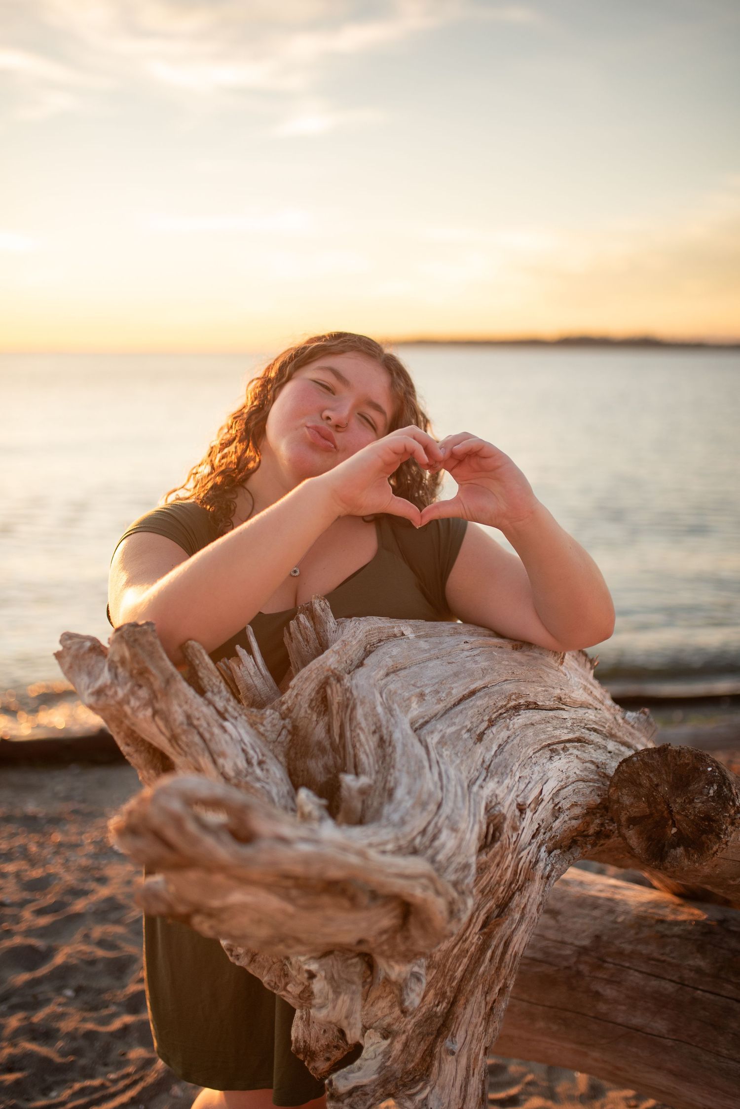 Young woman making heart shape with hands over driftwood log at beach during sunset.