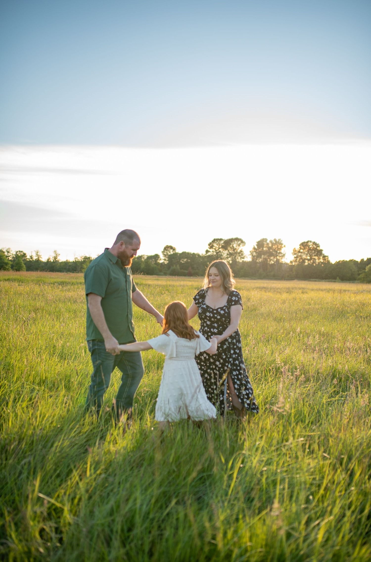 Family of three holding hands in golden sunlit meadow at sunset.