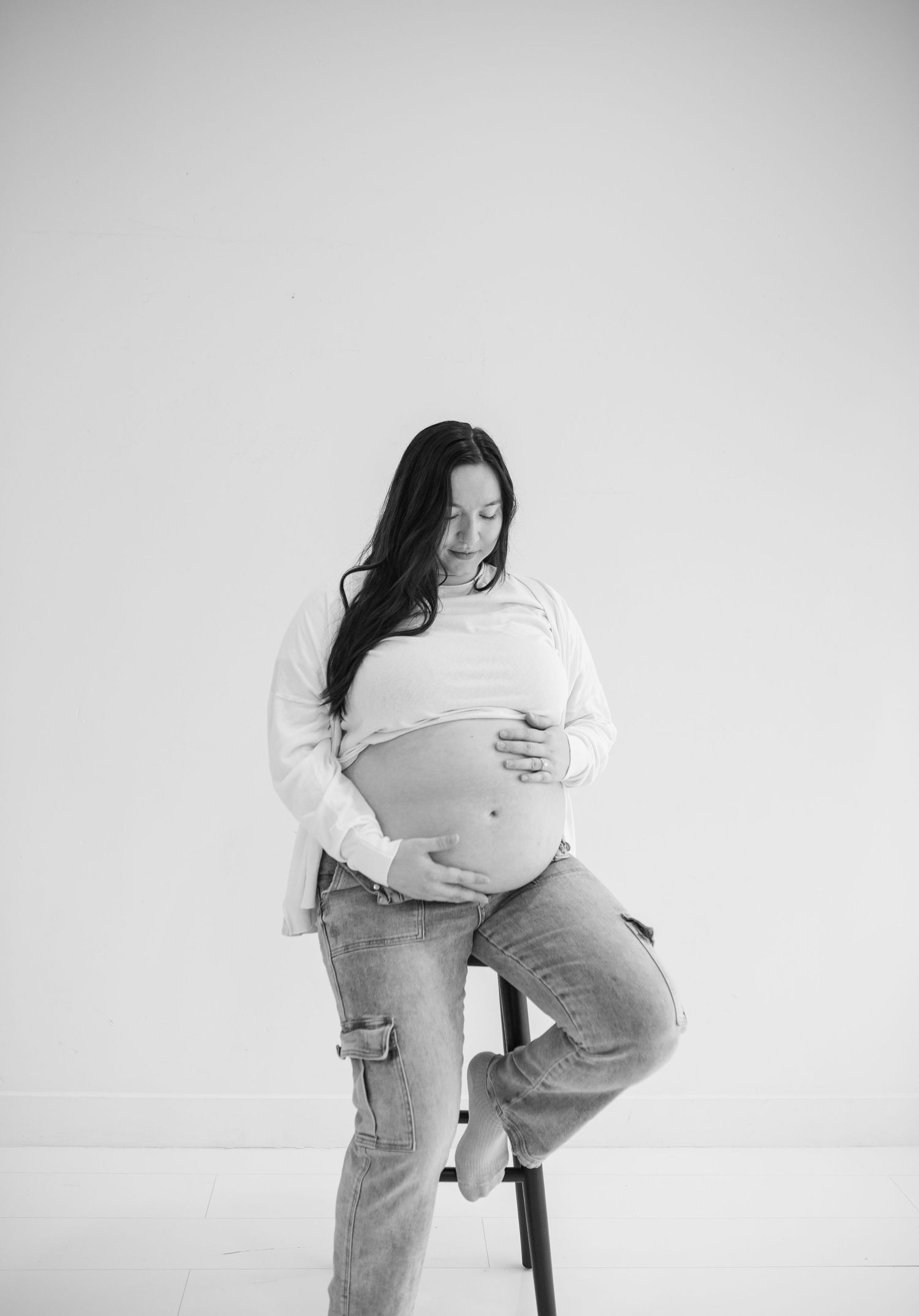 Black and white maternity portrait of pregnant woman sitting on stool looking down at her baby bump in white studio.