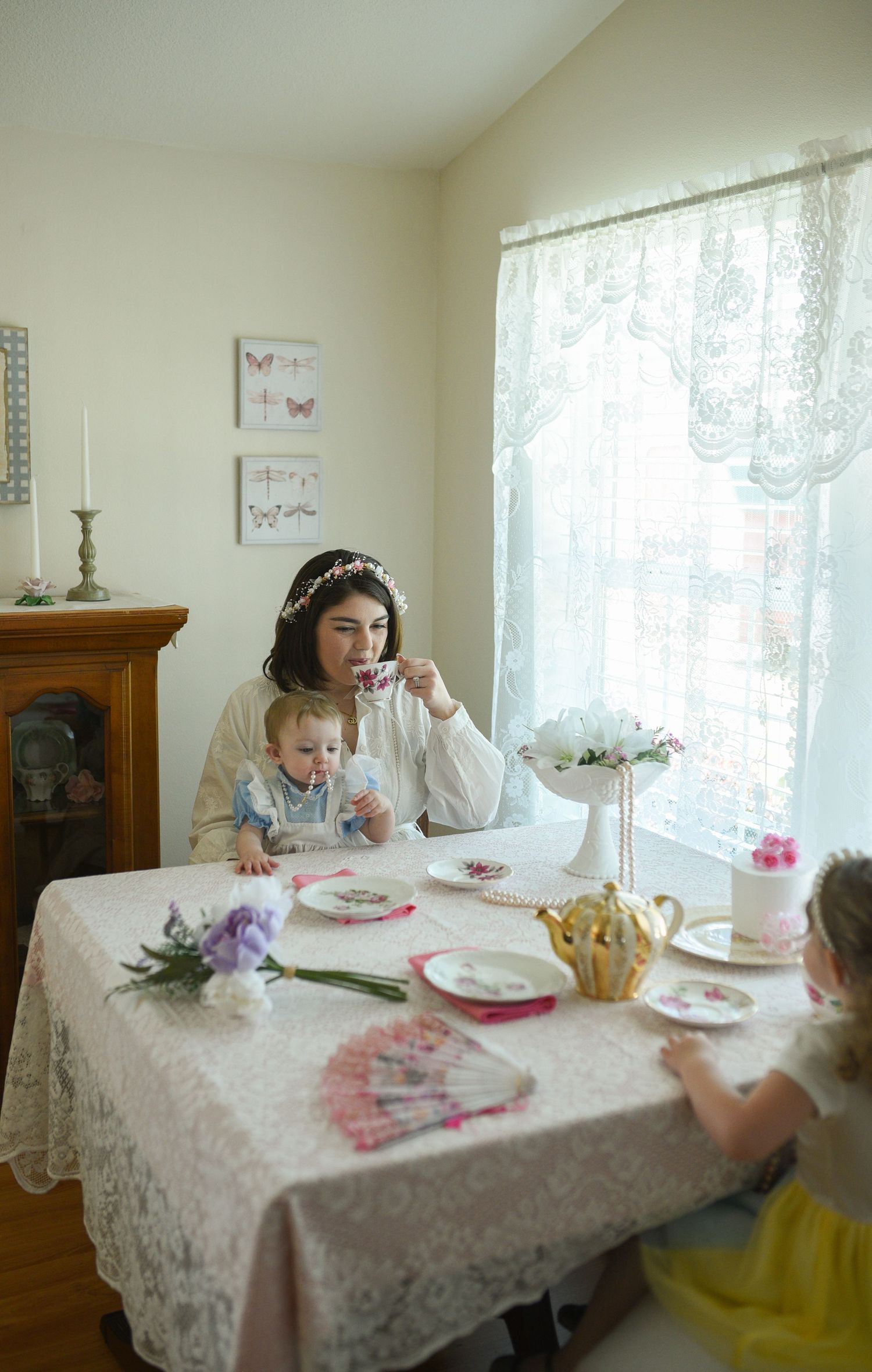 Woman in white dress sipping tea with two young children at a lace-covered tea party table.
