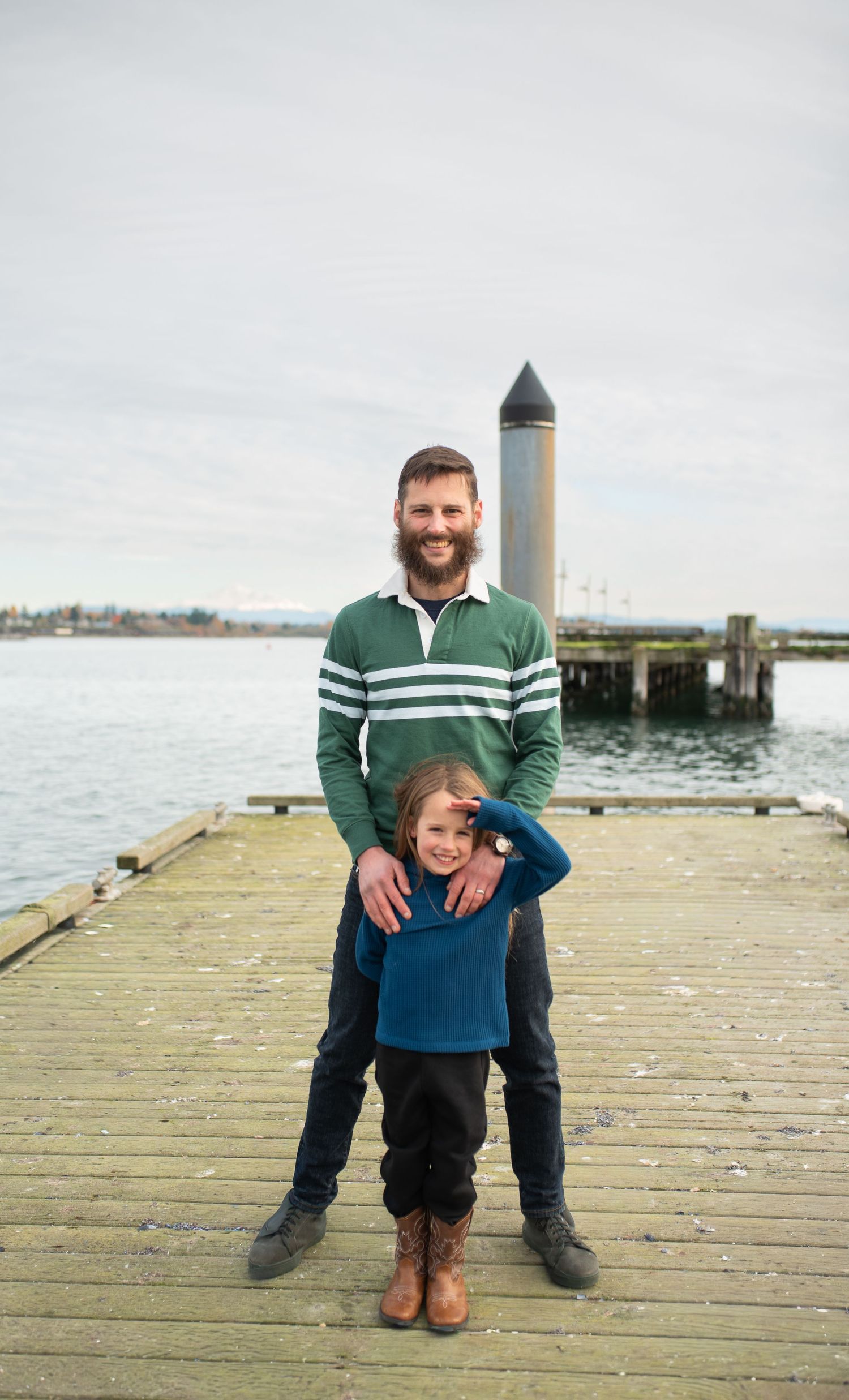 A smiling father stands on a waterfront dock with his young daughter in front of him.