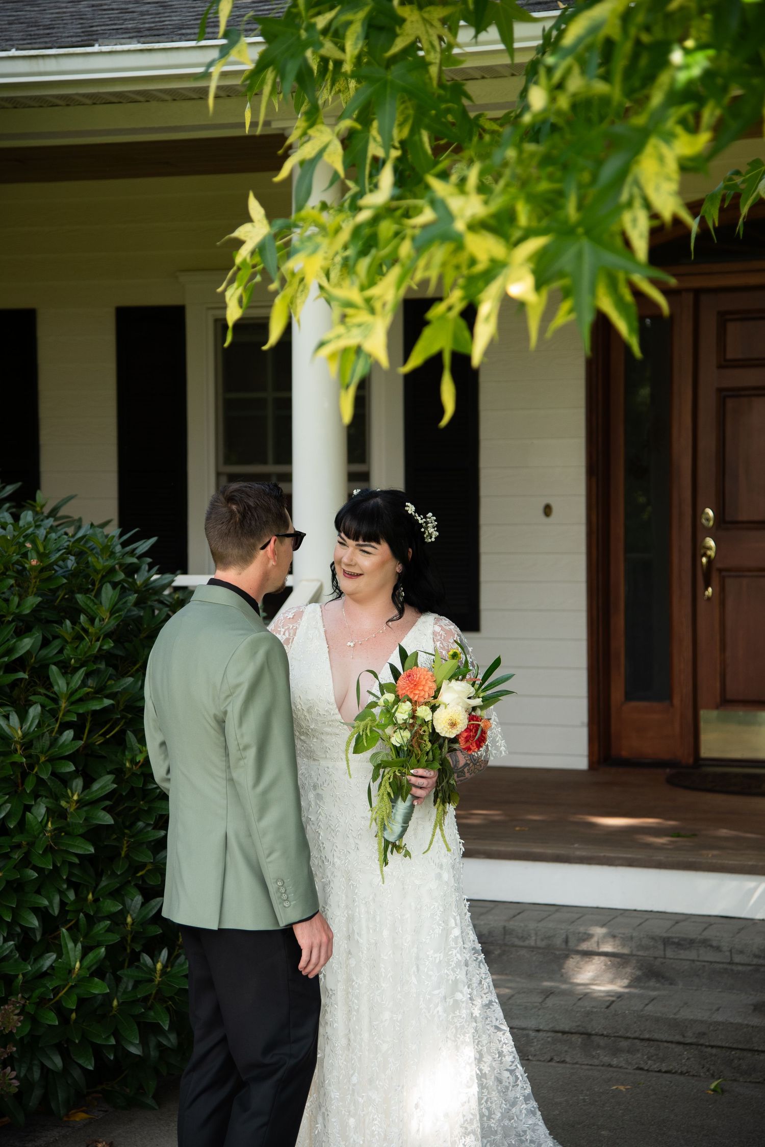 Bride in lace gown holds colorful bouquet while groom in sage green suit smiles at her outside white house.